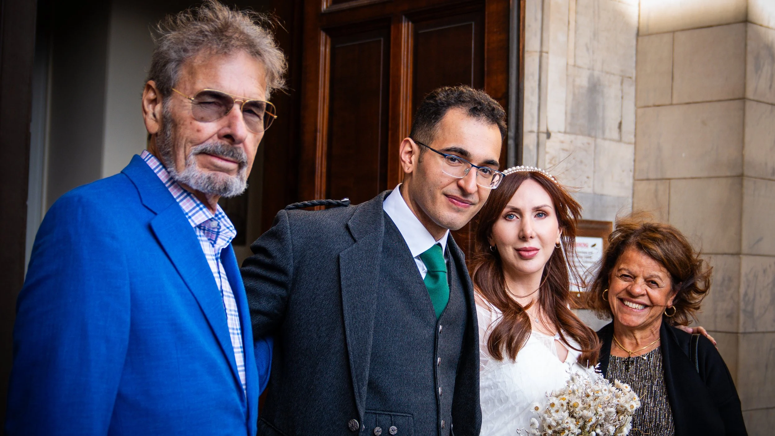 A group of four people, including a bride in white holding a bouquet, standing together for a photo inside a building with stone walls, during a wedding celebration.