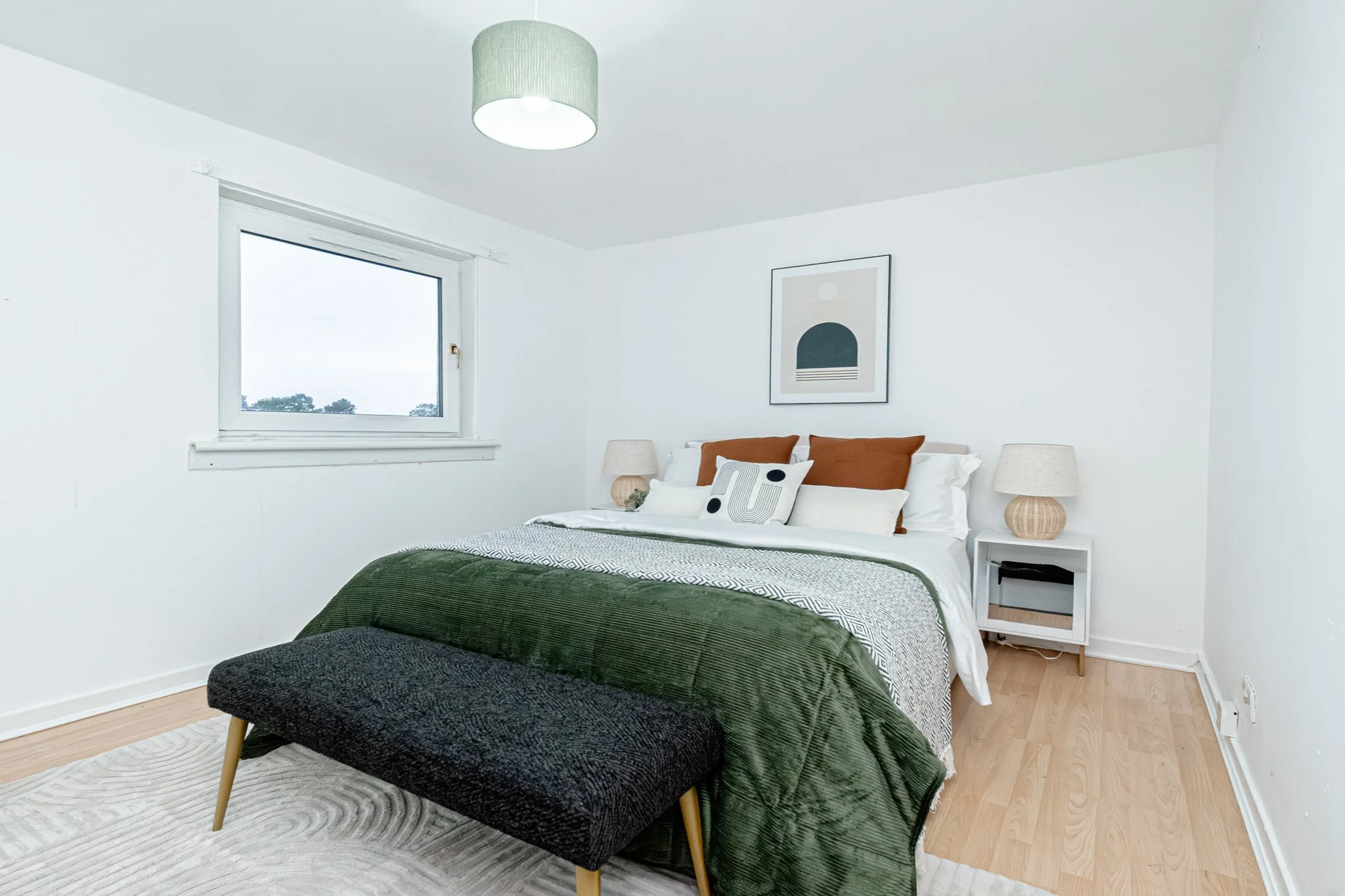 A bedroom with white walls, a bed with white and brown pillows, and a green velvet throw blanket. There are two white nightstands with beige lamps, a window with a view of trees, and a piece of abstract art on the wall.
