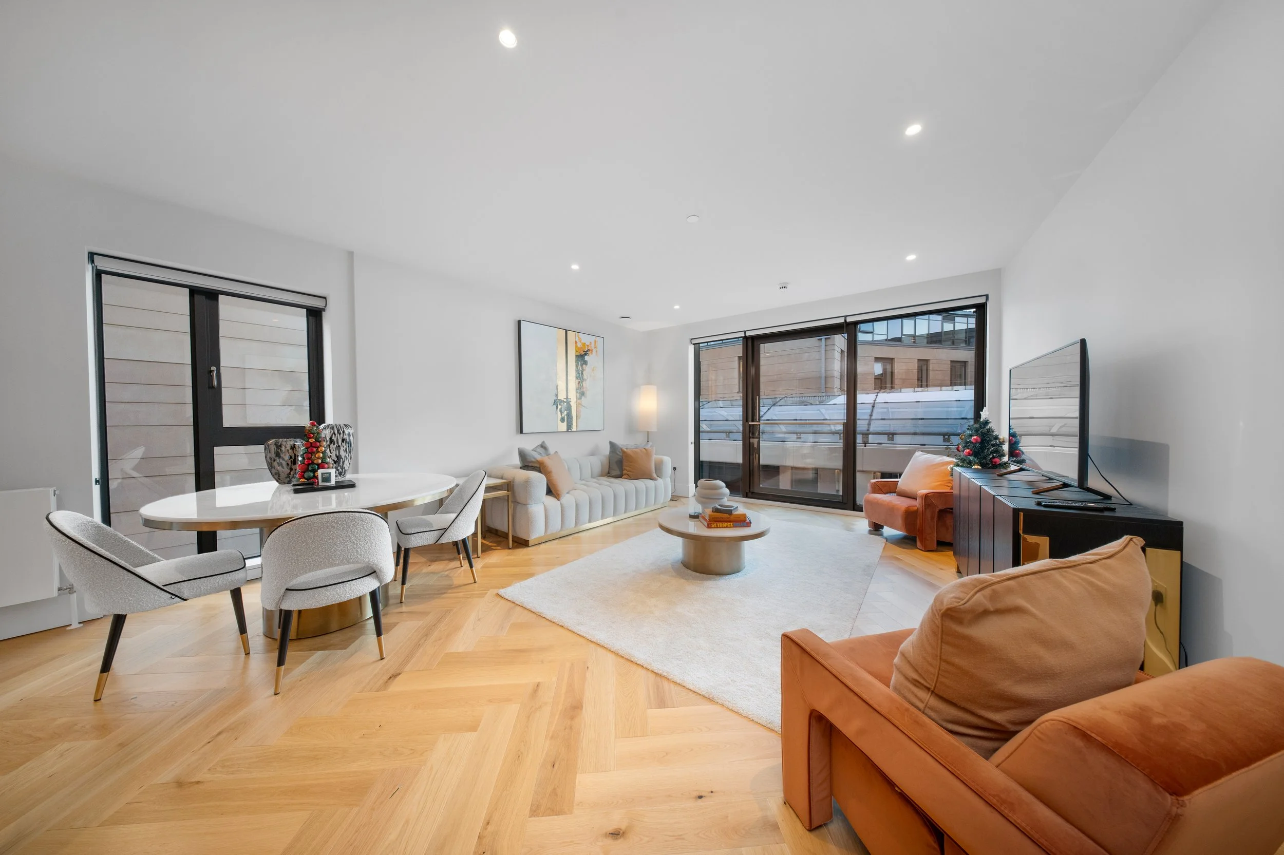 Modern living room with white walls, wooden flooring, a large window, beige sofa, orange armchair, round coffee table, and a flat-screen TV.