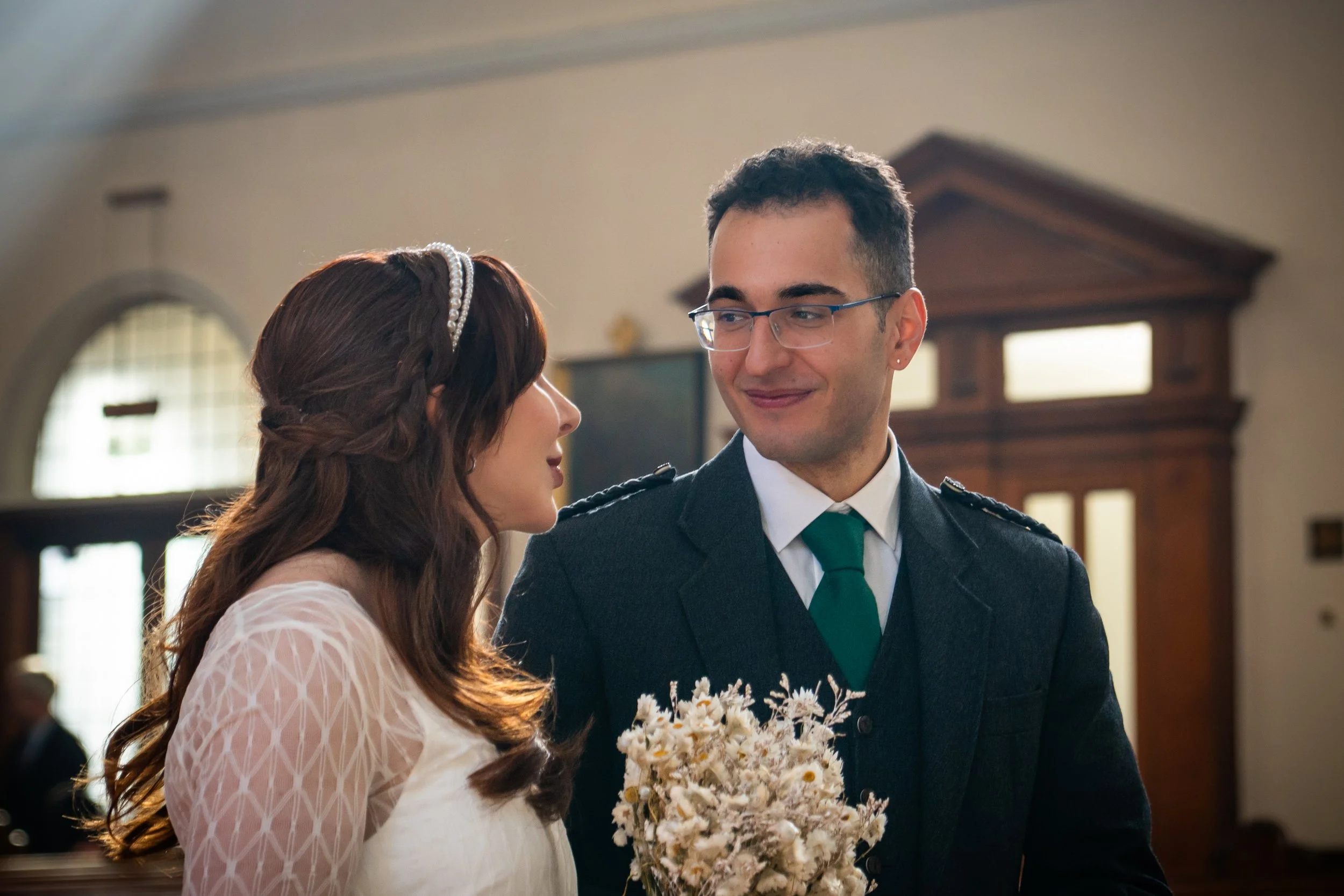 A bride and groom exchanging vows inside a church. The bride has long brown hair with a pearl headband, wearing a white lace dress and holding a bouquet. The groom has short dark hair, glasses, and is dressed in a dark suit with a green tie.