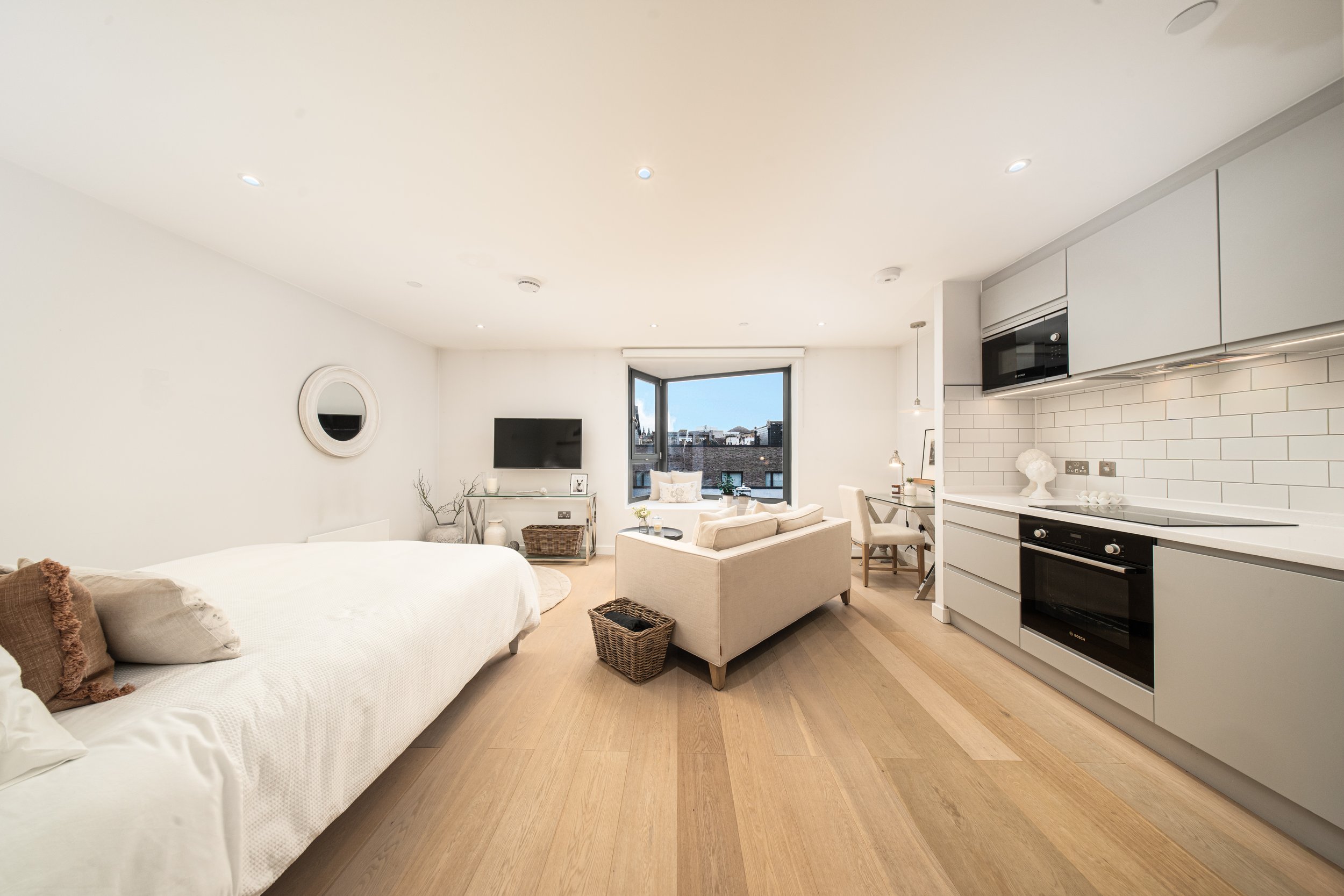 A modern, minimalist studio apartment with white walls, light wood flooring, a bed on the left, a small sitting area with a sofa and chairs near the window, a wall-mounted TV. Real Estate Edinburgh