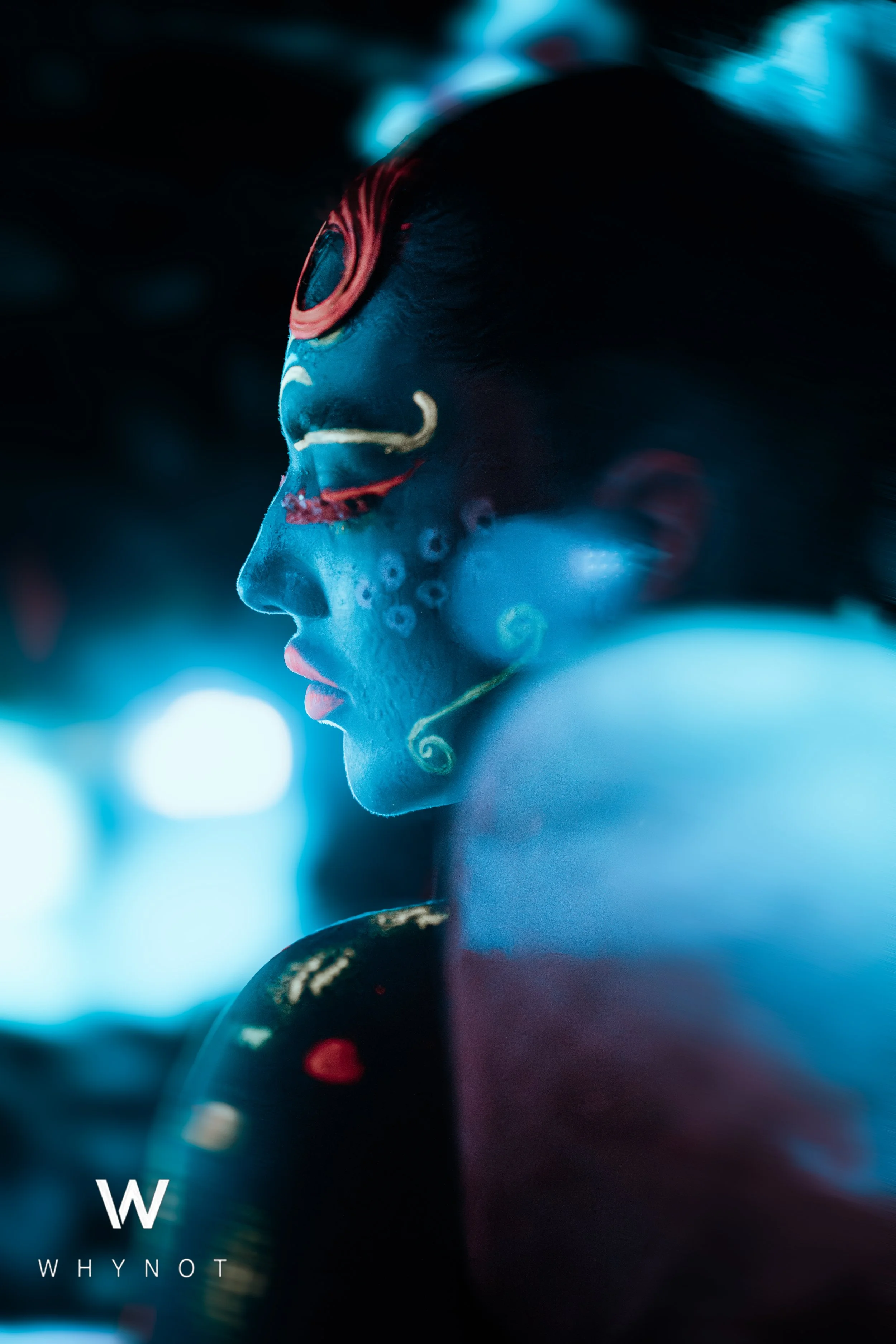 A woman with face painted in vibrant colors, illuminated by blue and red lighting, with intricate patterns and designs on her face.