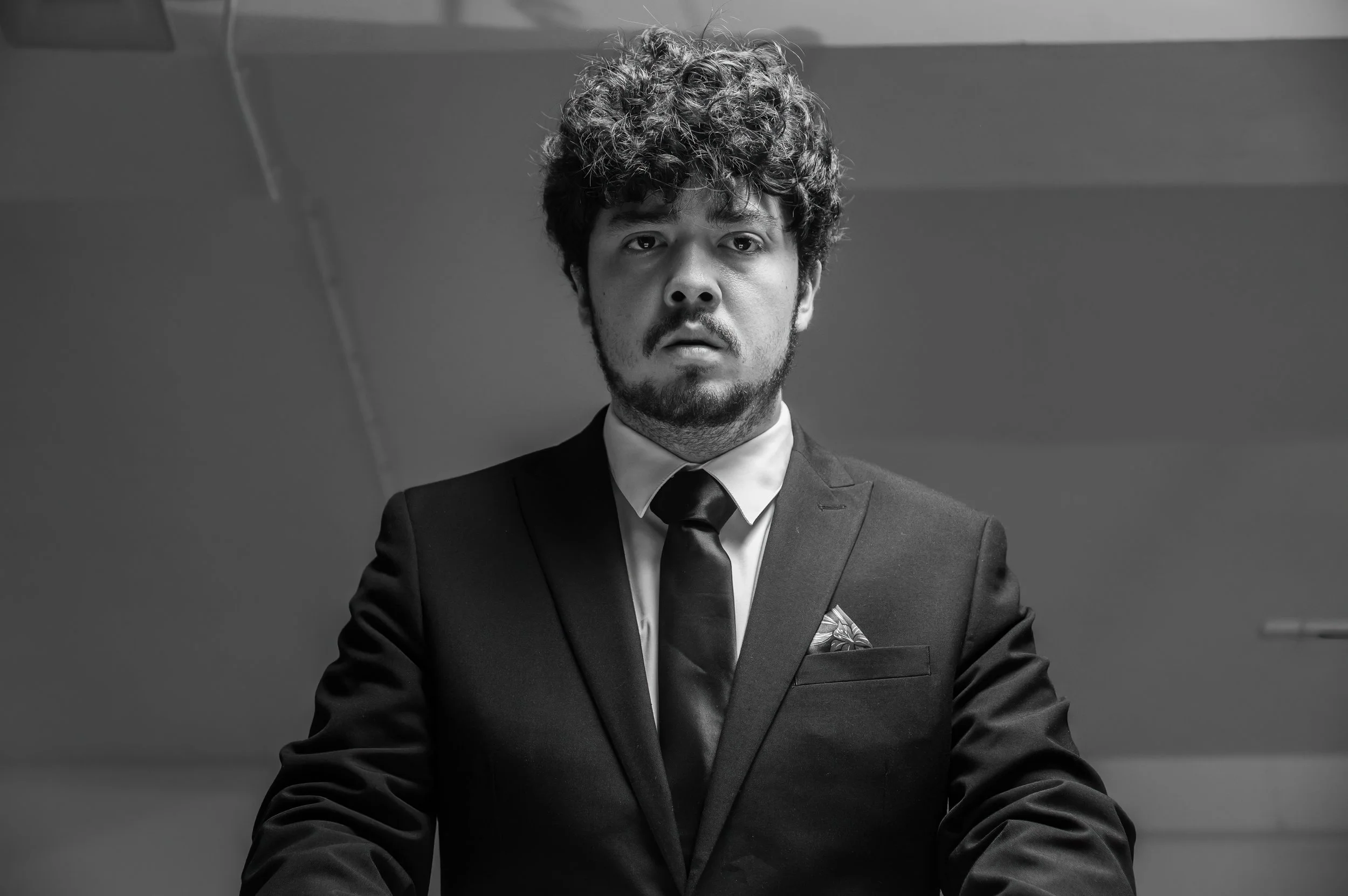 Black and white close-up photo of a man with curly hair and a beard wearing a suit, tie, and pocket square, looking directly at the camera with a serious expression.