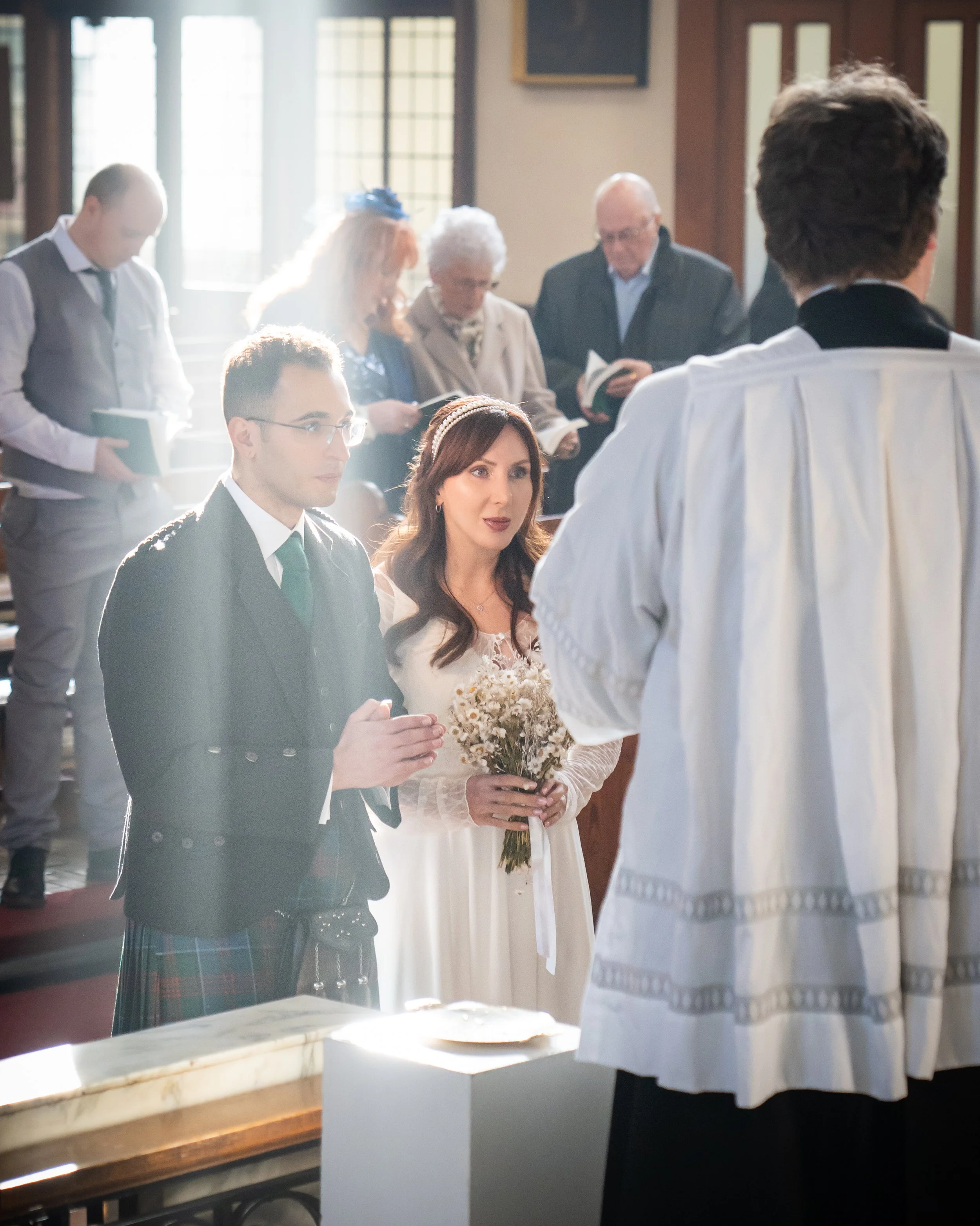 A wedding ceremony in a church with a bride holding a bouquet and a groom in a suit, standing before a priest during the vows, with guests in the background.