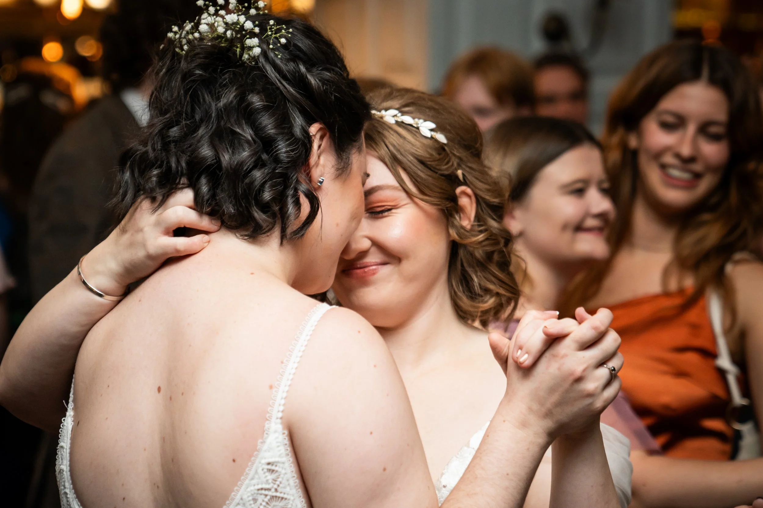 Two women, likely at a wedding, are hugging and smiling with their eyes closed, surrounded by other women who are also smiling and enjoying the moment.