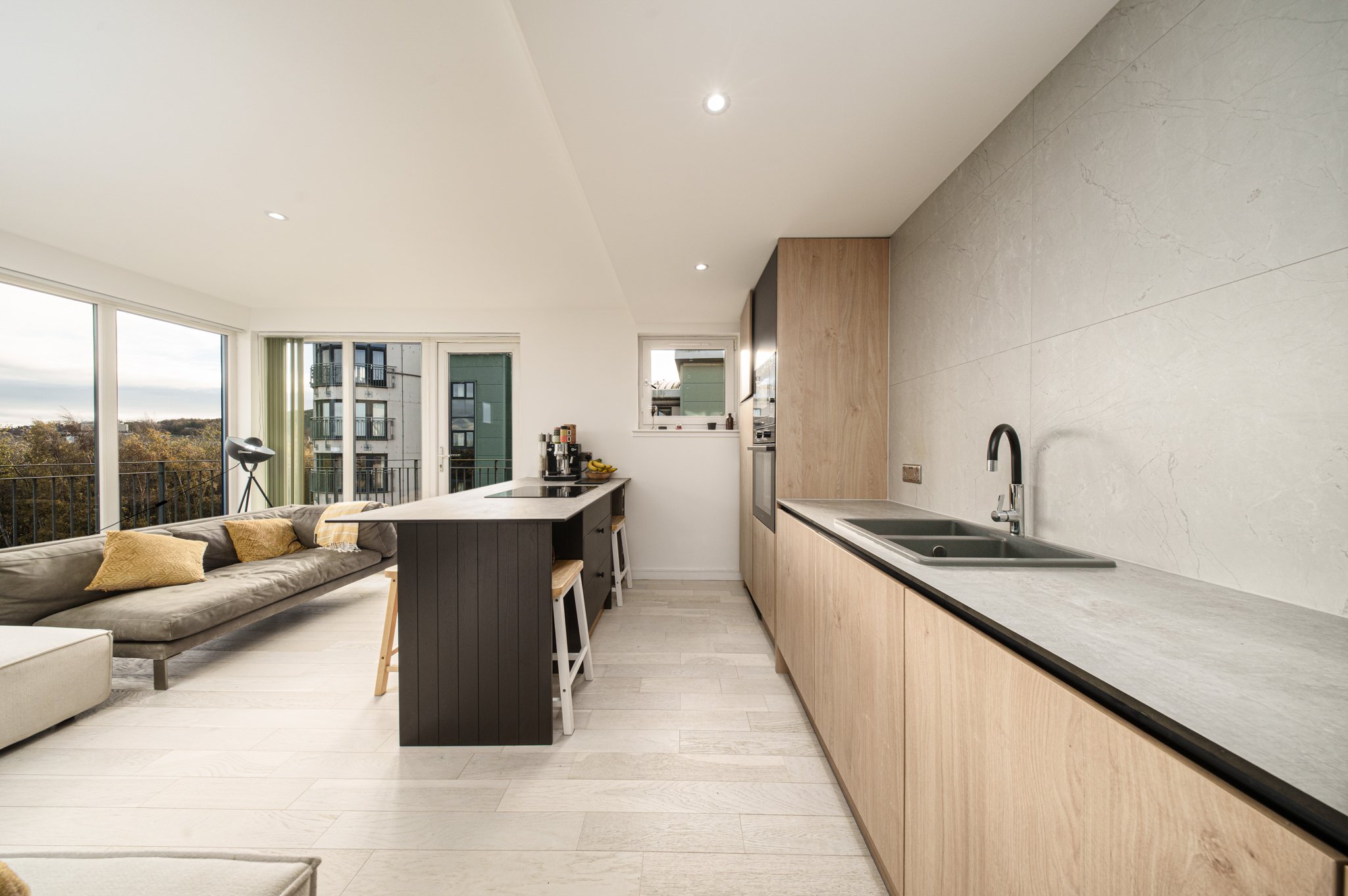 Modern open-concept kitchen and living room with large windows, a beige sofa with pillows, a kitchen island with bar stools, countertop appliances, and wood cabinetry.