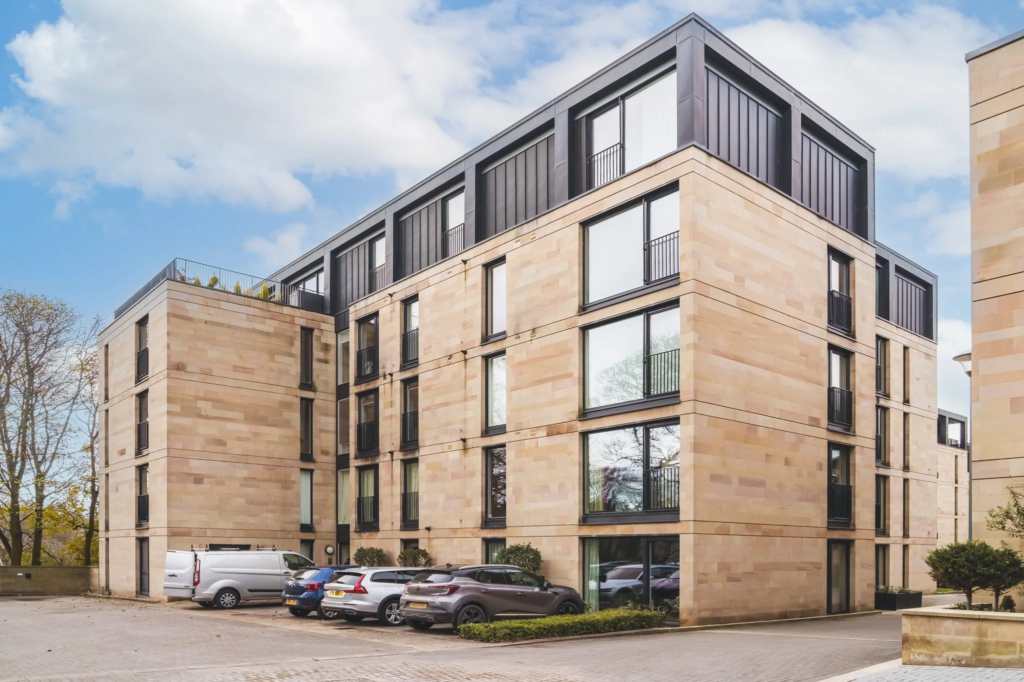 Modern five-story apartment building with beige stone exterior and large windows, parking lot with several cars, trees in background, partly cloudy sky. Real Estate Edinburgh