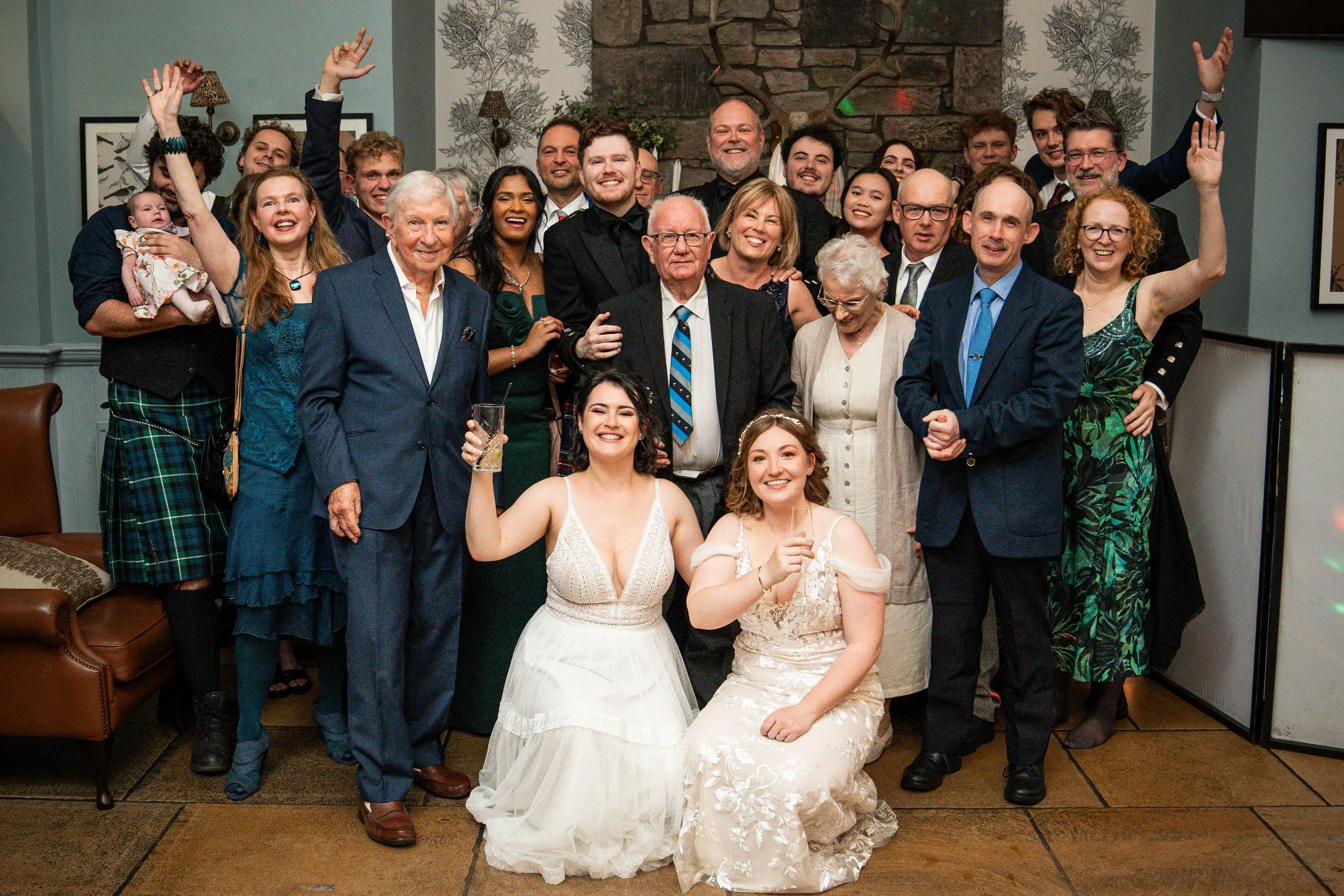A large group of people gathered indoors at a celebration, with some dressed in formal attire and others in casual or traditional clothing. Two women in white dresses are seated in front, holding drinks, smiling. The group appears joyful, with many smiling and some raising their hands.
