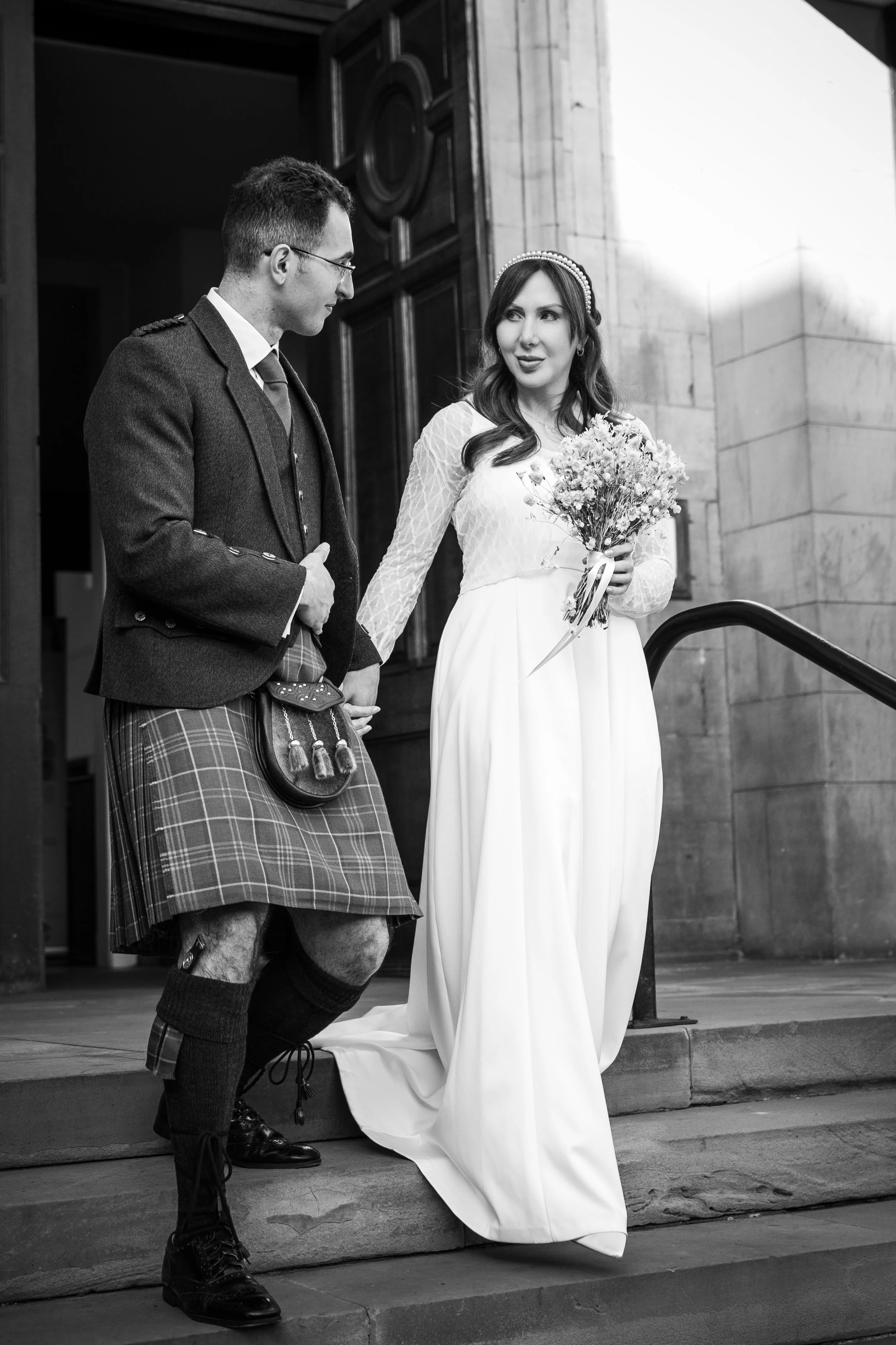 A black and white photo of a bride and groom walking down steps. The bride is holding a bouquet of flowers and wearing a long white dress and a headband. The groom is dressed in traditional Scottish attire, including a kilt, jacket, and sporran, holding the bride's hand as they descend the stairs outside a stone building.
