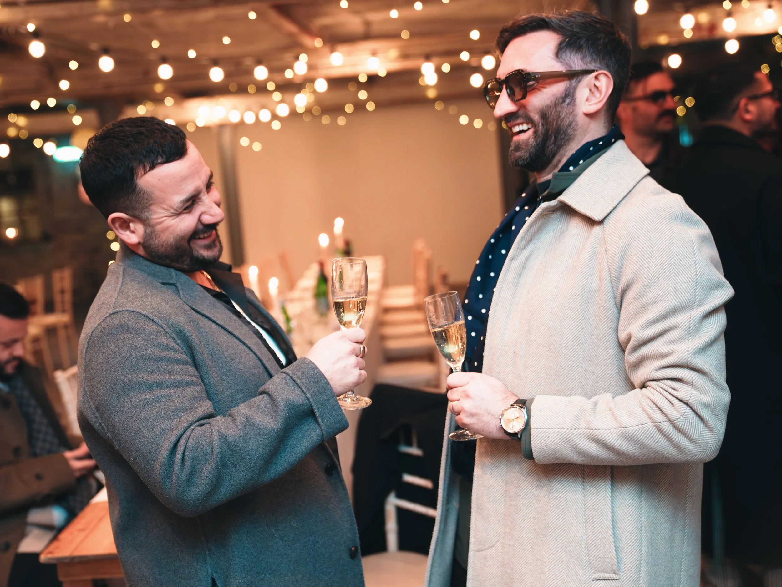 Two men in suits smiling and holding glasses of champagne at a party or celebration under warm string lights.