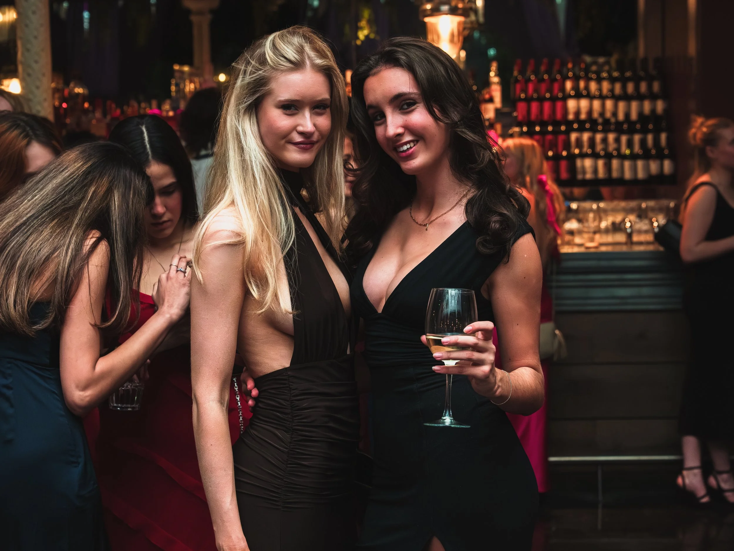 Two women in elegant black dresses at a social event, one holding a glass of white wine, in a lively, dimly-lit bar or club setting with other people in the background.