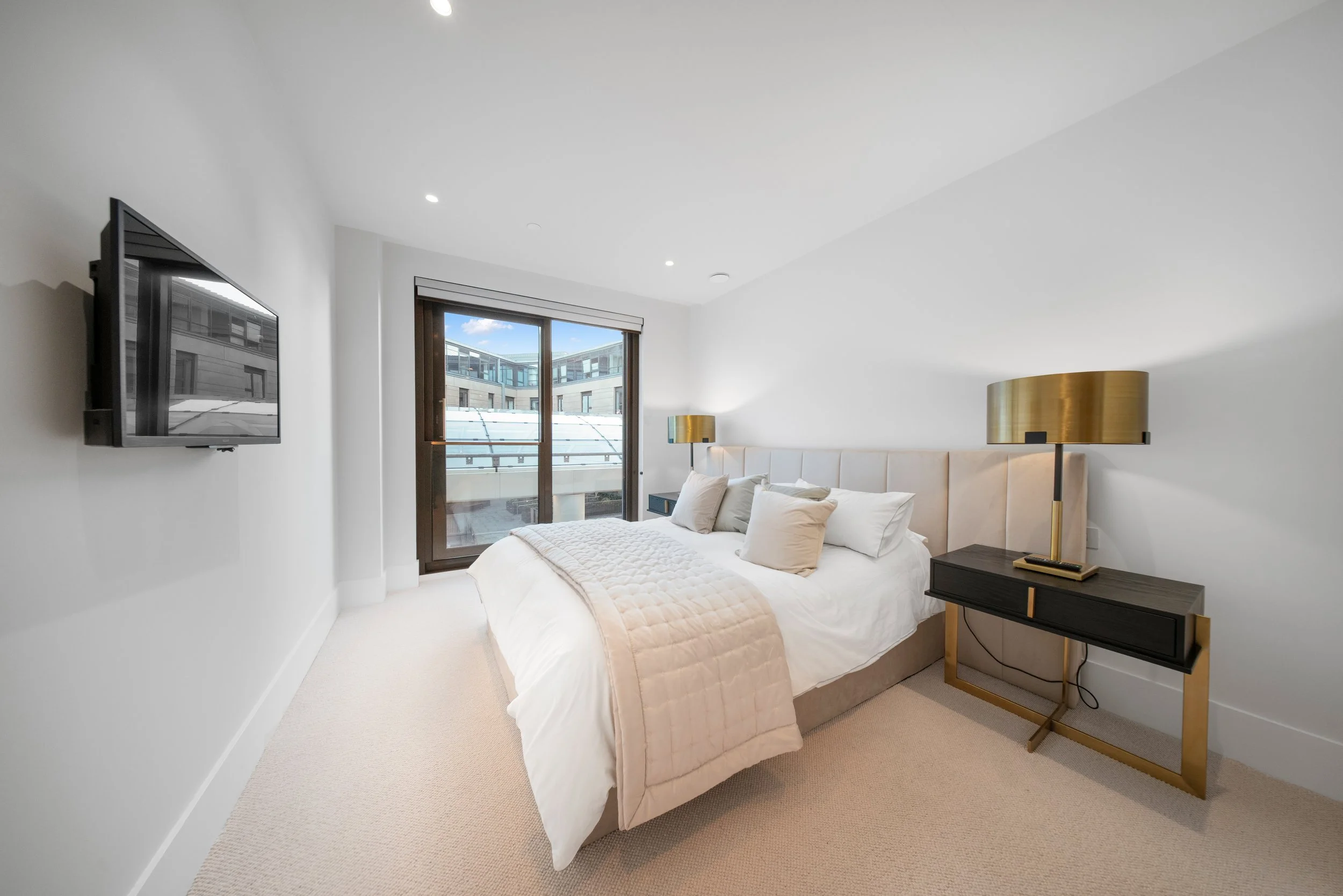 Modern bedroom with a large bed, white bedding, beige headboard, two gold and black lamps, black nightstand, balcony doors with a view of an apartment complex, and a wall-mounted TV Real Estate Edinburgh