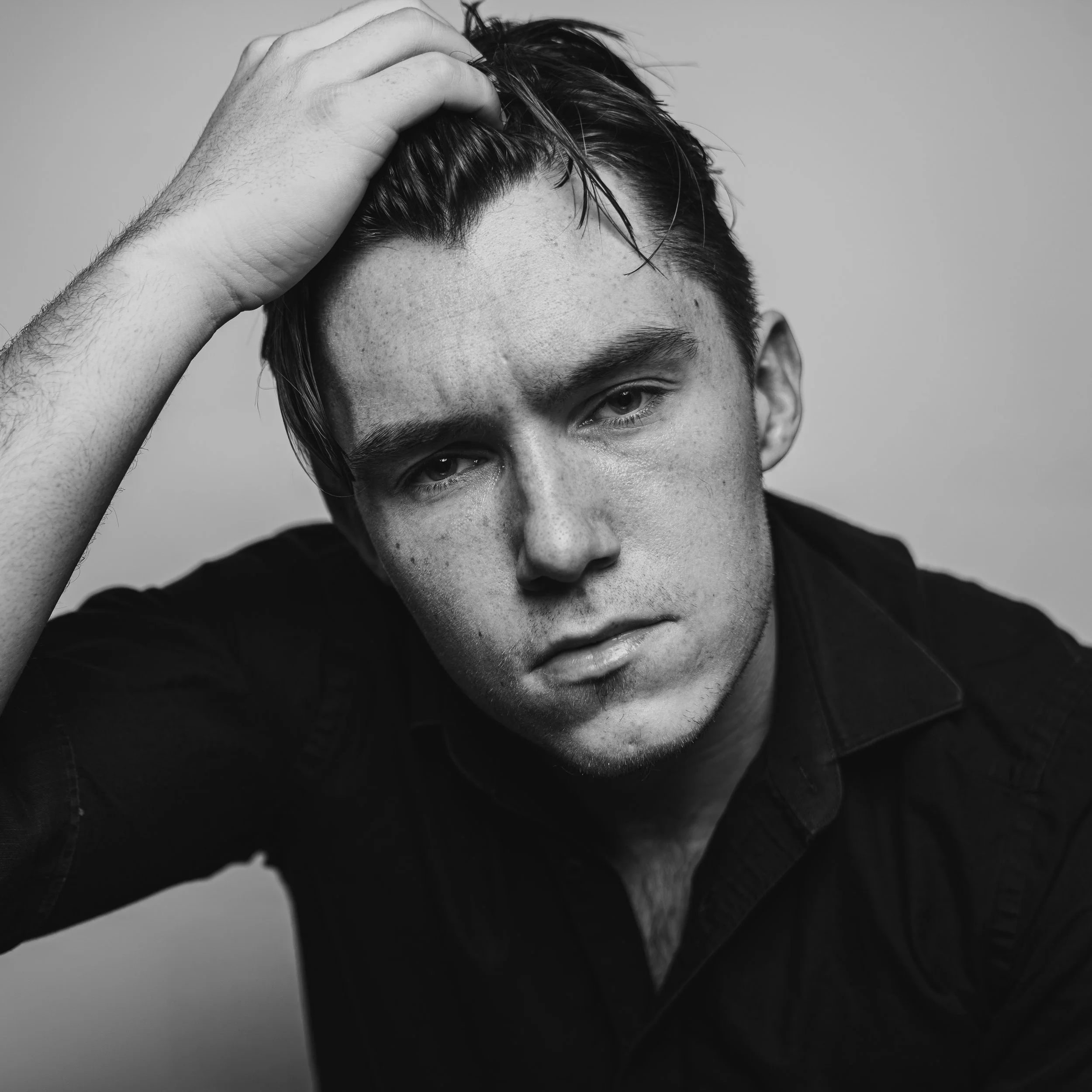 Black and white portrait of a young man with short dark hair, resting his hand on his head, wearing a dark shirt, with a serious expression.
