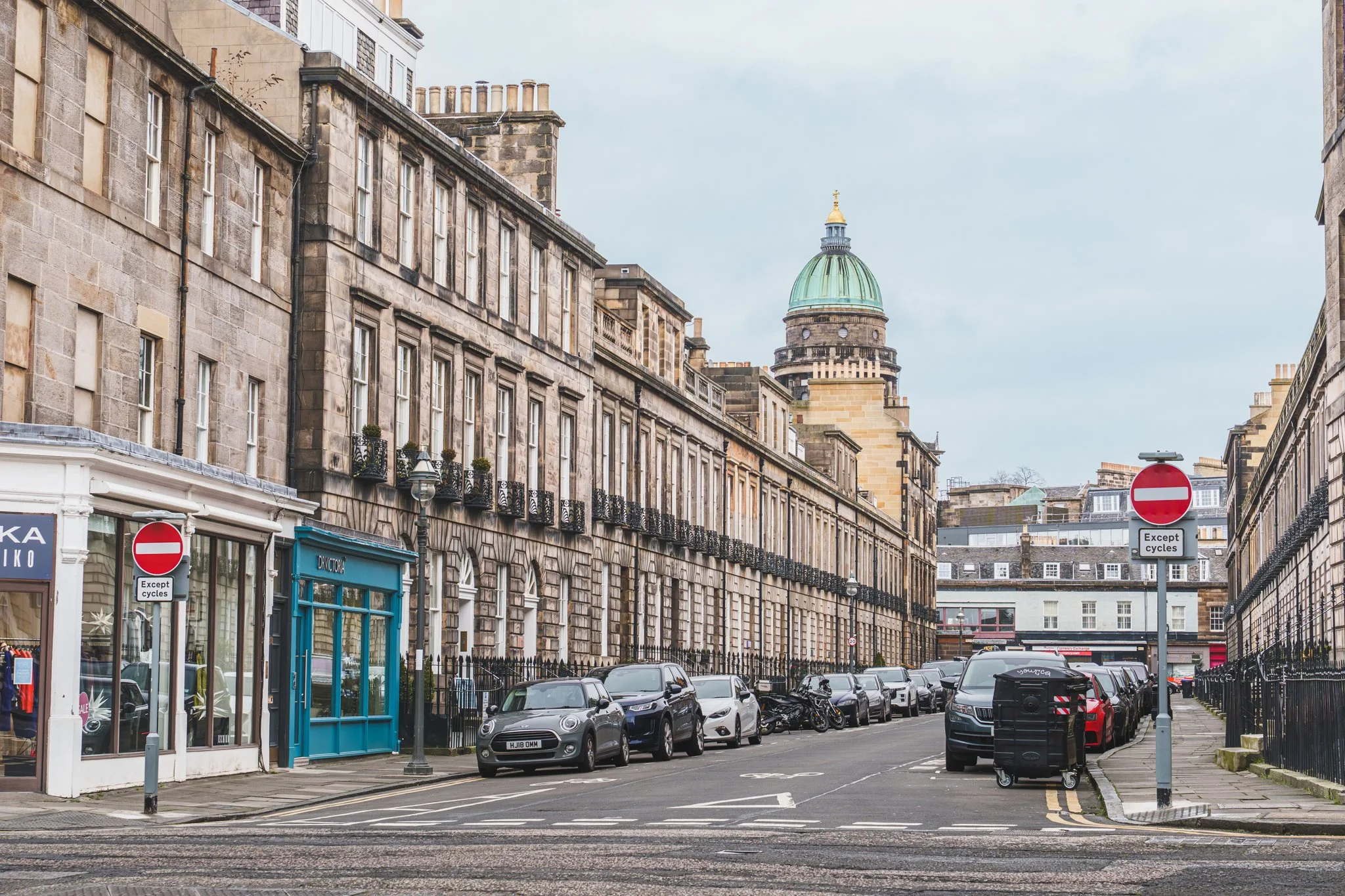A city street with parked cars along stone buildings, a blue storefront, and a green-domed tower in the background.