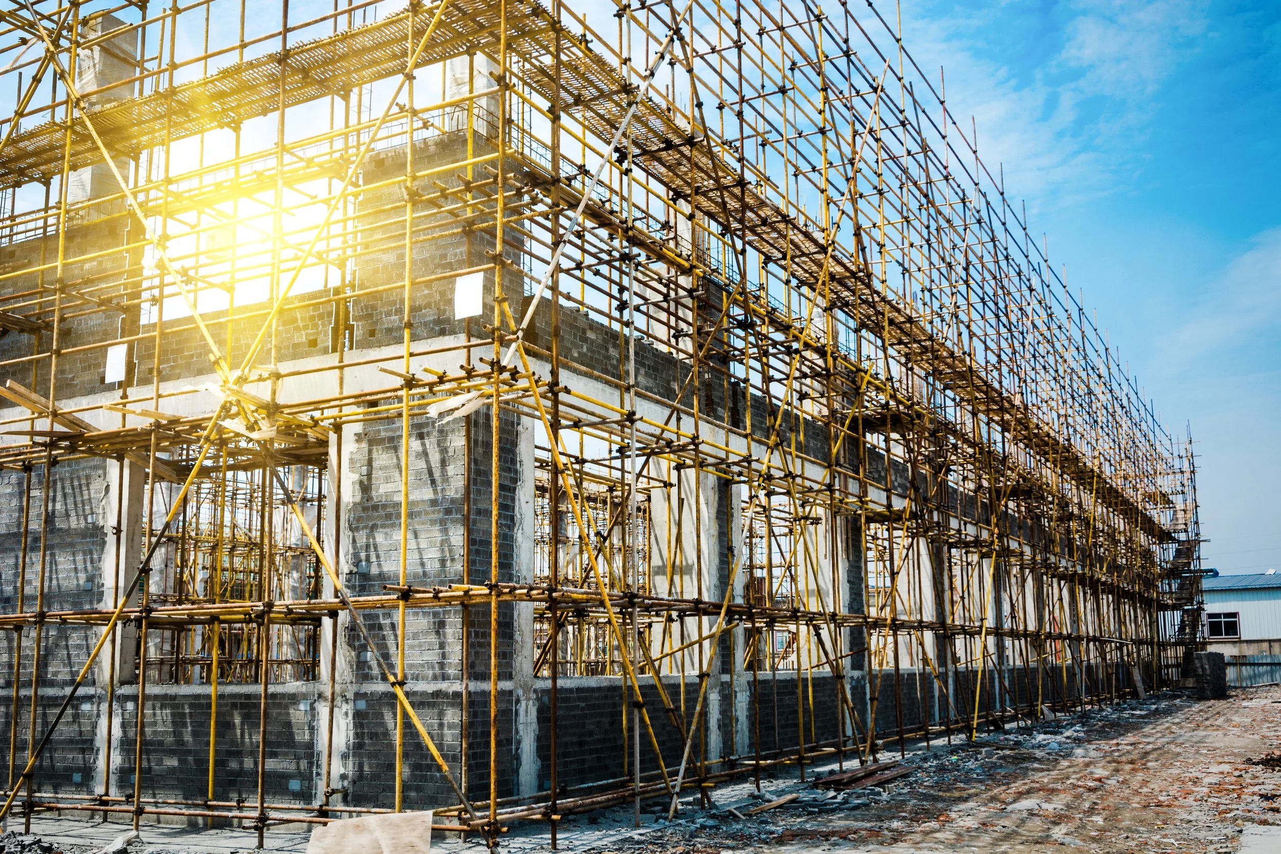 Construction site featuring a building under construction with yellow scaffolding and brick walls, sunlight shining through, and a blue sky in the background.