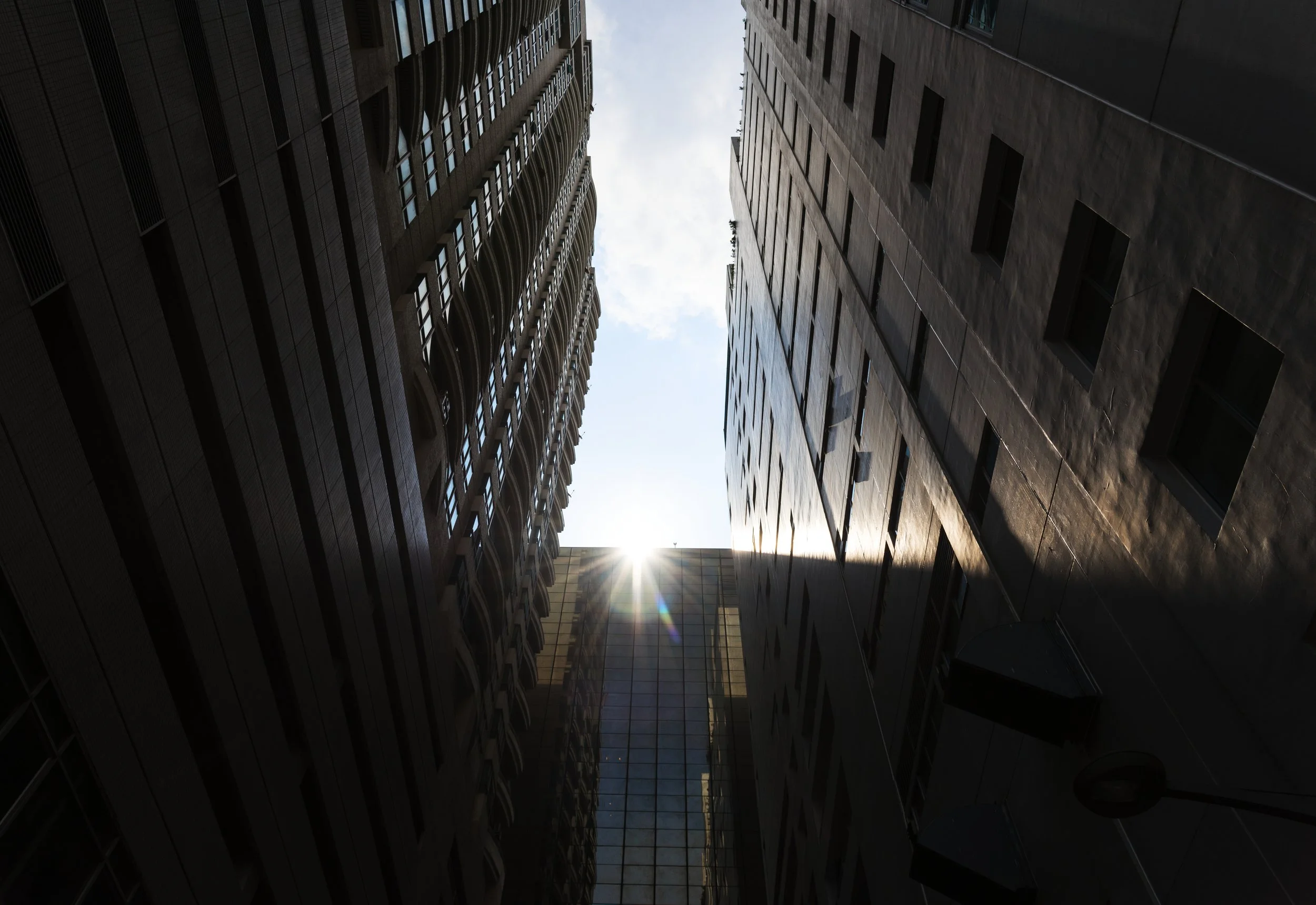 Looking up between tall skyscrapers with the sun shining between them.