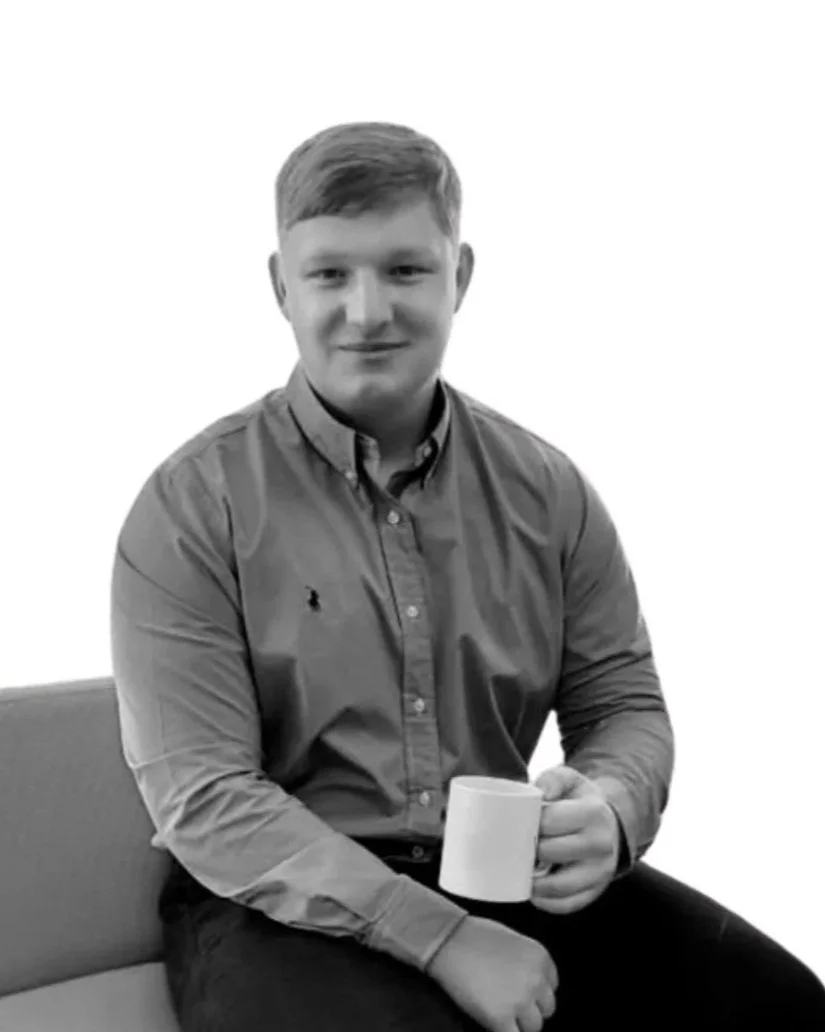 A person sitting indoors on a couch holding a mug, with a plant beside them and a window in the background.