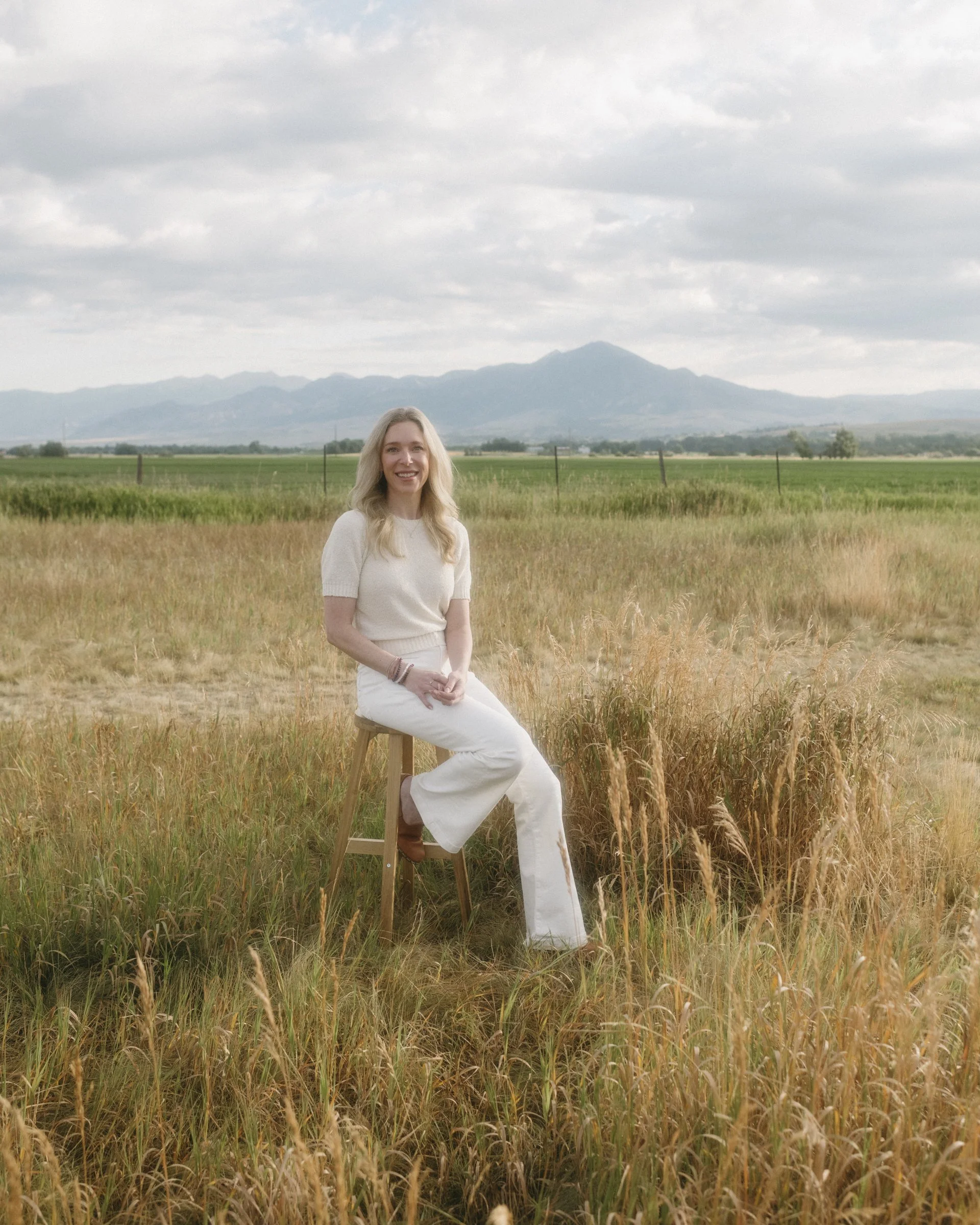 A woman with long blonde hair, dressed in white, sitting on a stool in a golden field of tall grass with a mountain range and cloudy sky in the background.
