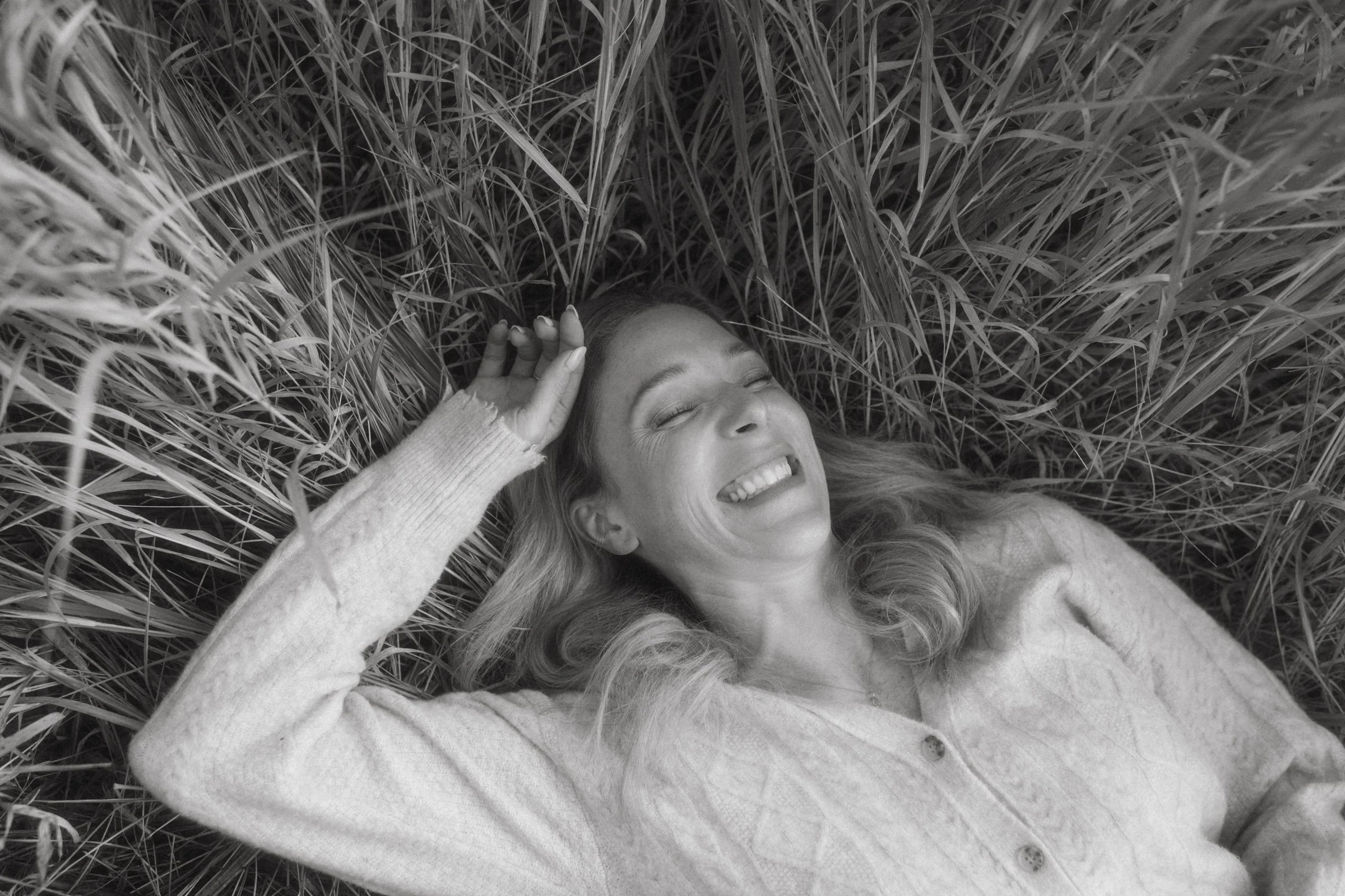 A woman with long hair lying on her back in tall dry grass, smiling with eyes closed and her hand resting on her forehead.