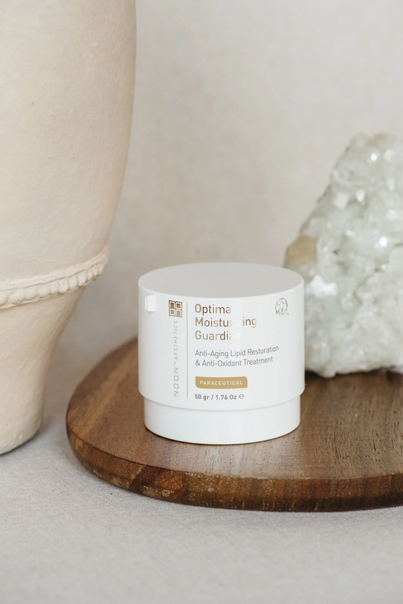 A white container labeled 'Optima Moisturizing Guardian' sitting on a round wooden platform, surrounded by beige and off-white textured objects and rocks.