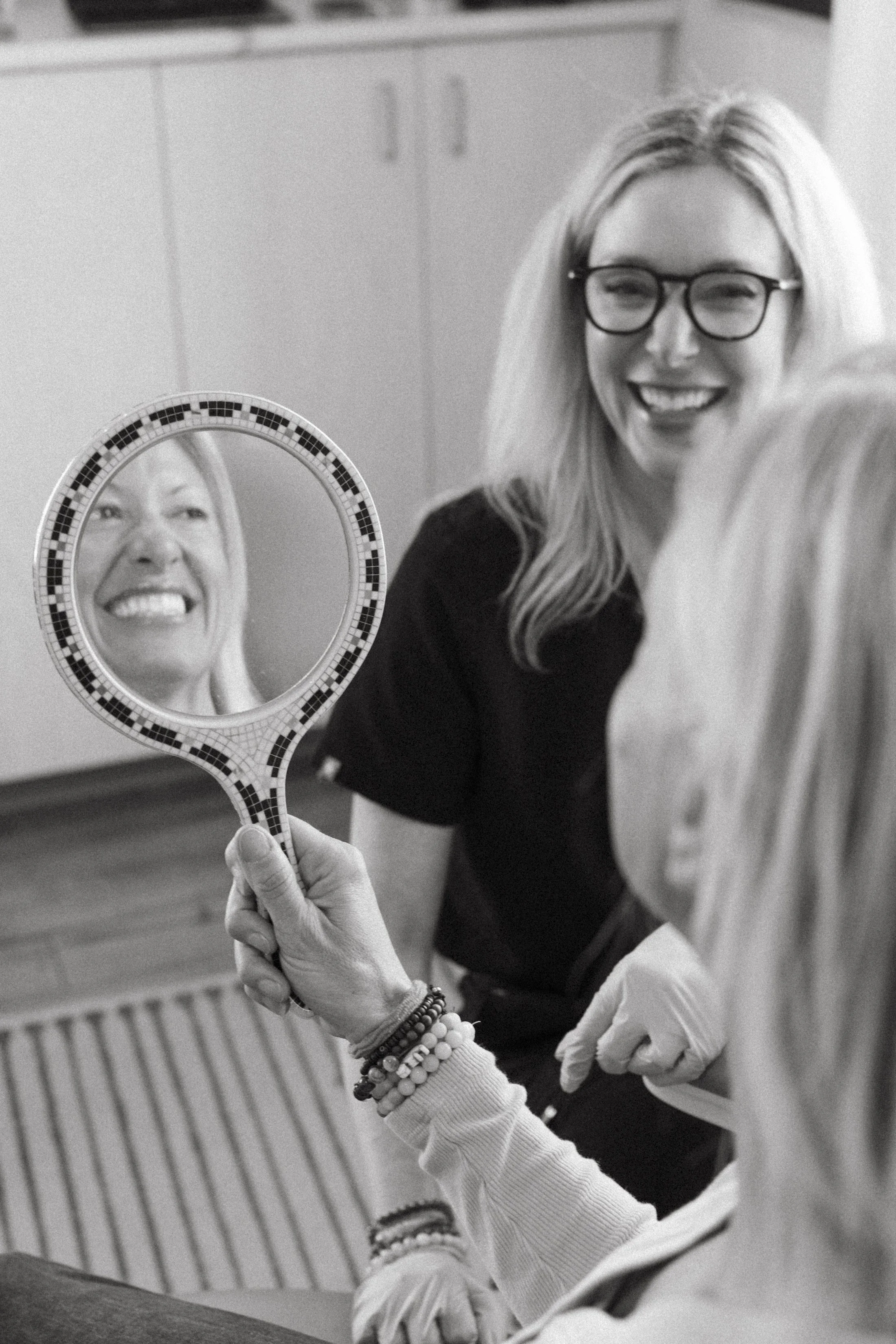 Woman with glasses smiling, holding a mirror, wearing multiple bracelets, seen from the shoulders up in black and white.