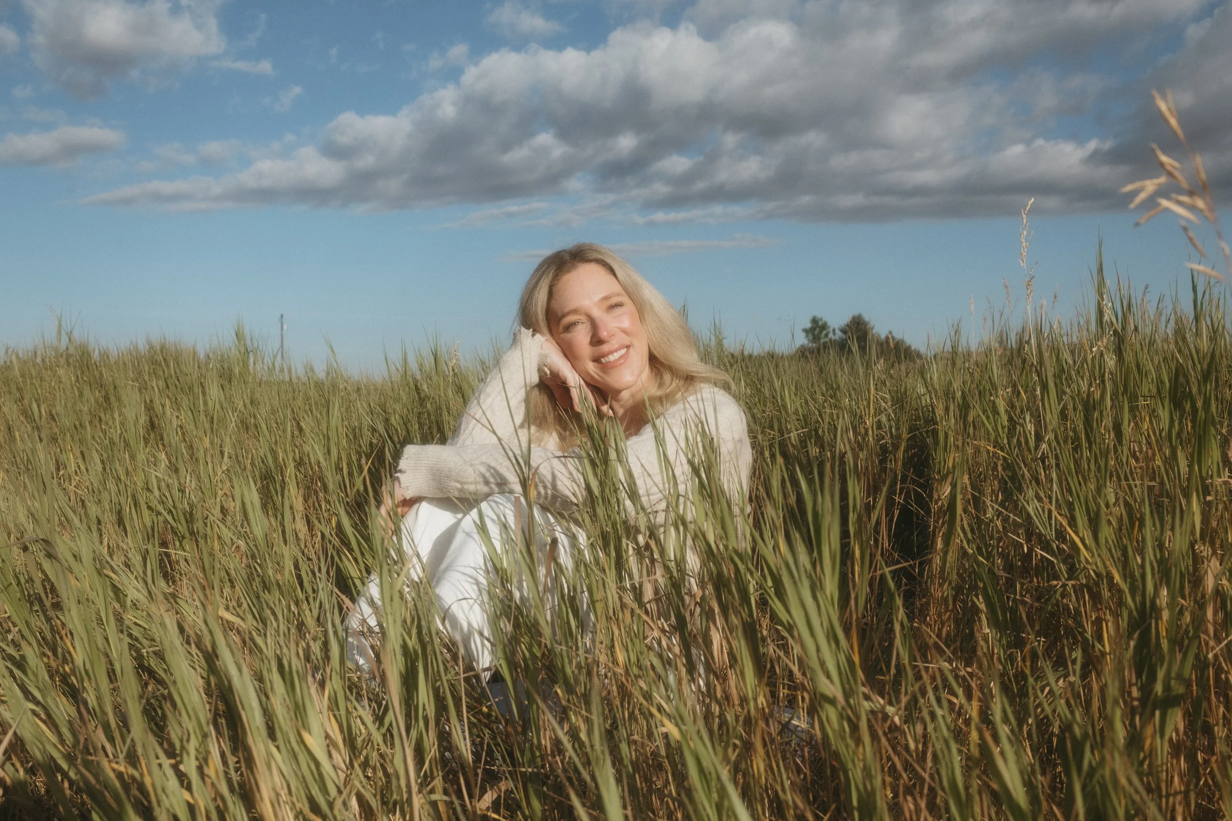 A woman with blonde hair smiling and resting her head on her hand while sitting in tall grass in a field, under a cloudy sky with blue patches.
