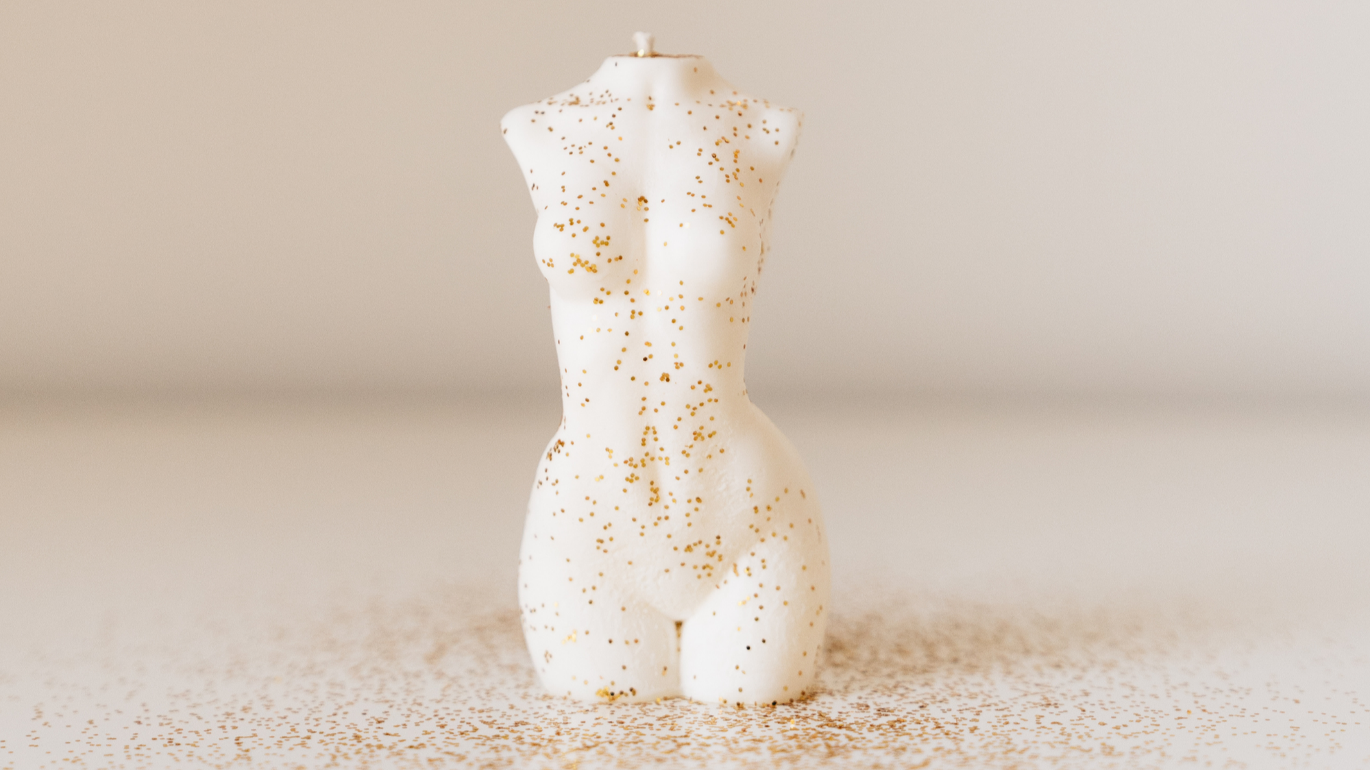 A sculpted white candle in the shape of a female torso, decorated with scattered gold glitter and placed on a light surface with a neutral background.