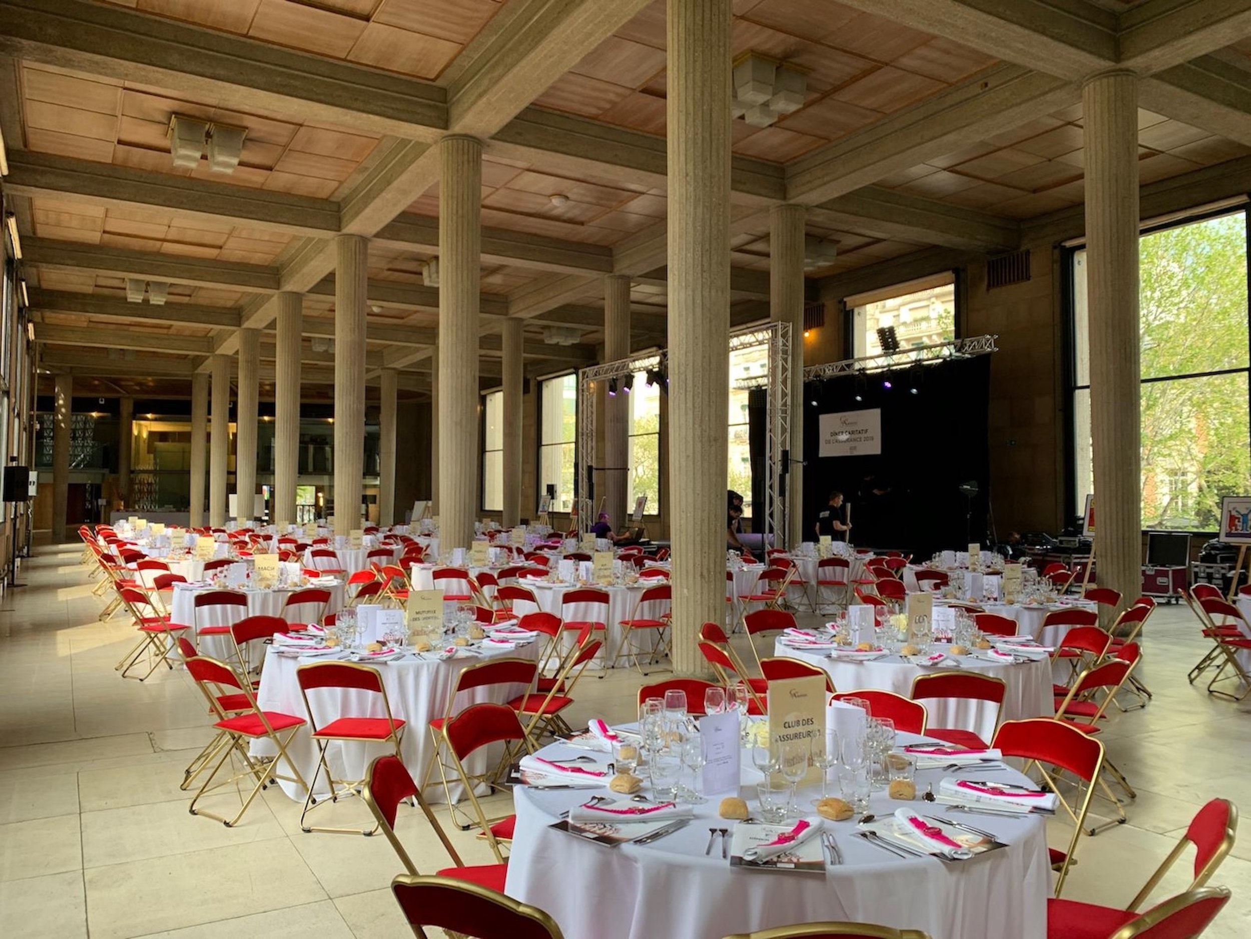 Grande salle avec des tables rondes dressées, nappes blanches, chaises rouges, et plafonds hauts avec colonnes.