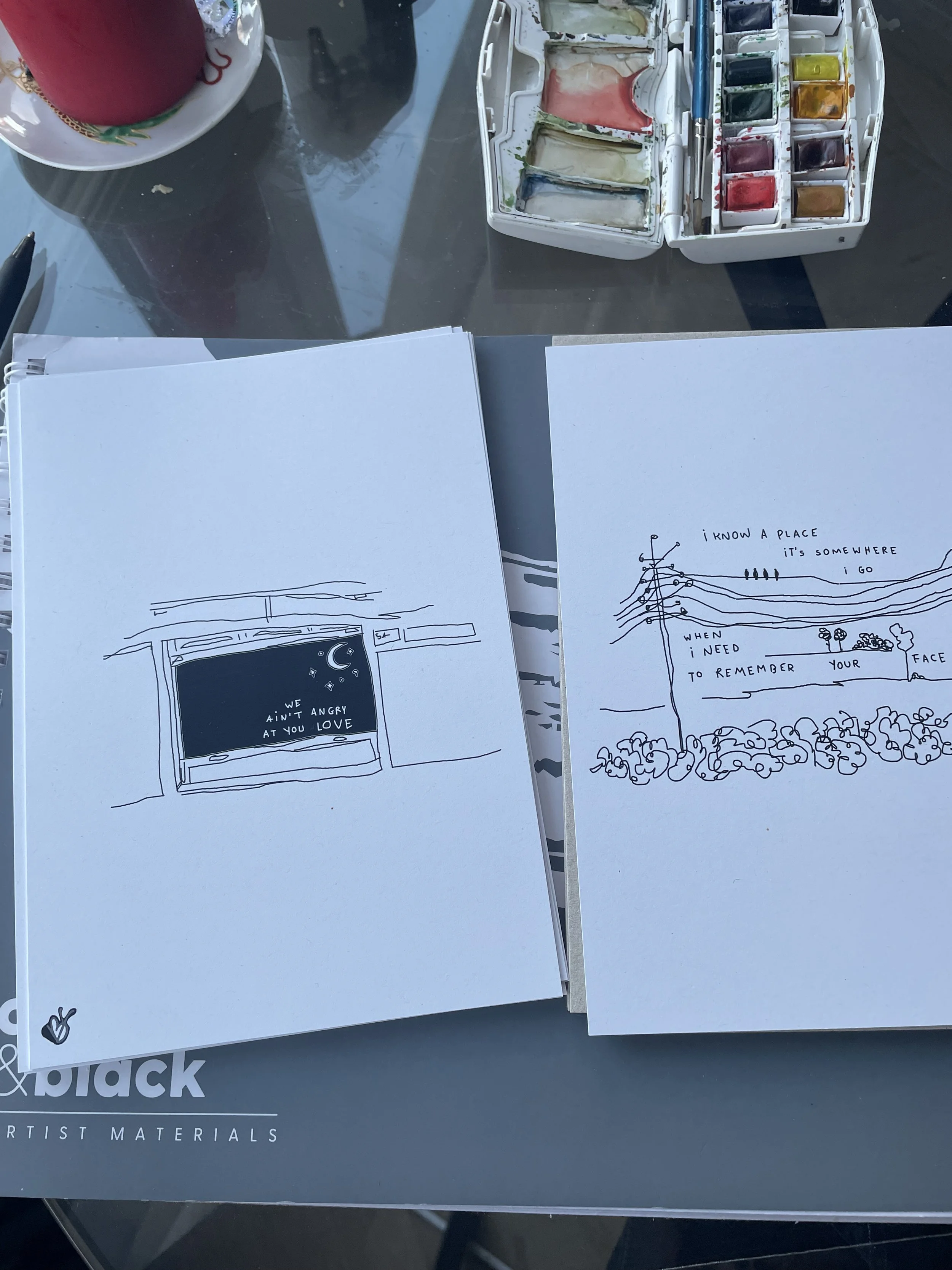Two sheets of paper with black ink sketches and handwritten text, situated on an artist's workspace. The left sheet depicts a window with stars and the words 'We ain't angry at you love.' The right sheet shows a landscape with power lines and trees, 