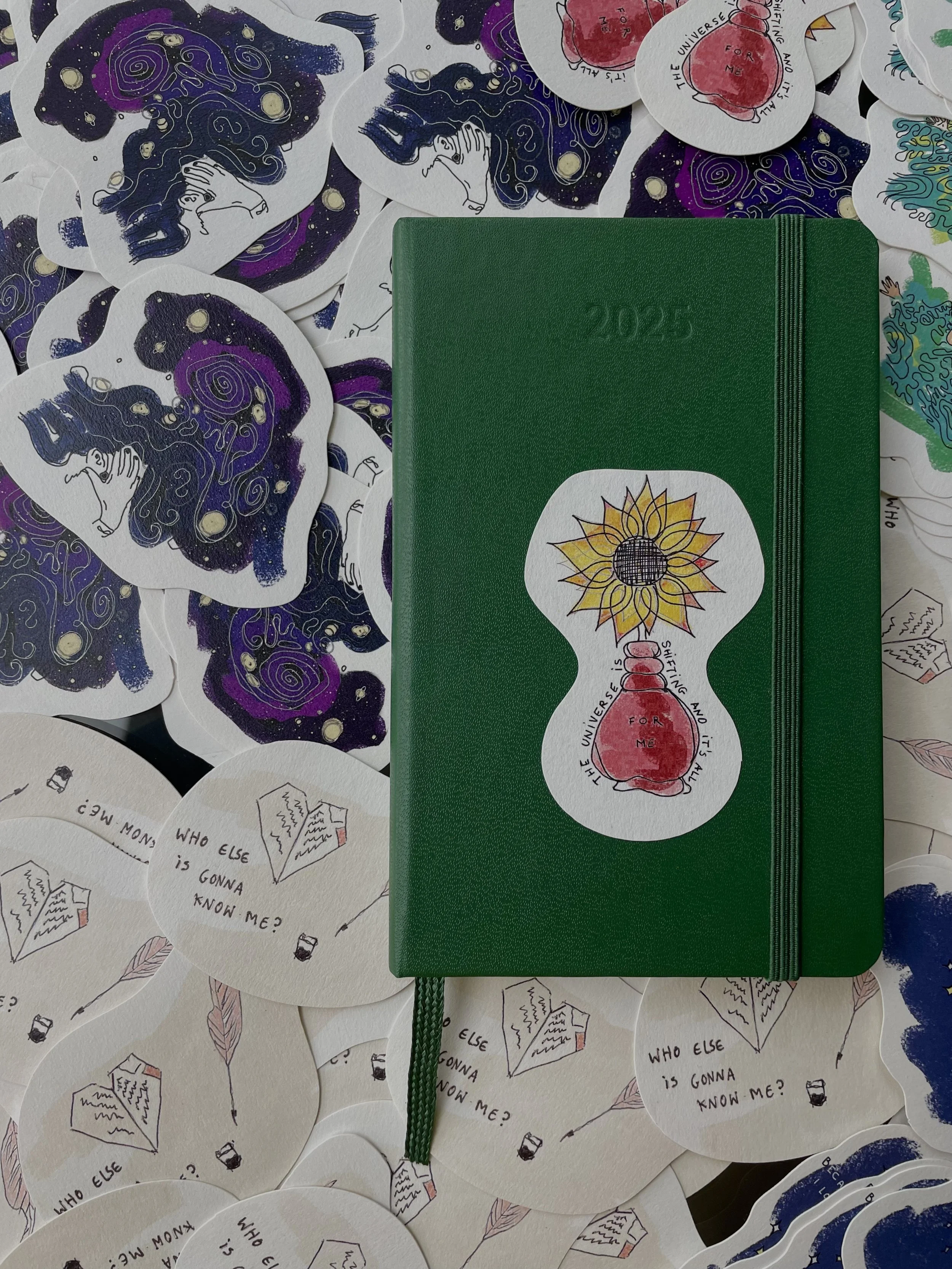 A green 2025 planner with a sticker of a sunflower and a red heart on the cover, surrounded by various hand-drawn stickers including cosmic scenes, hearts, and books with the phrase "Who else is gonna know me?" written on some.
