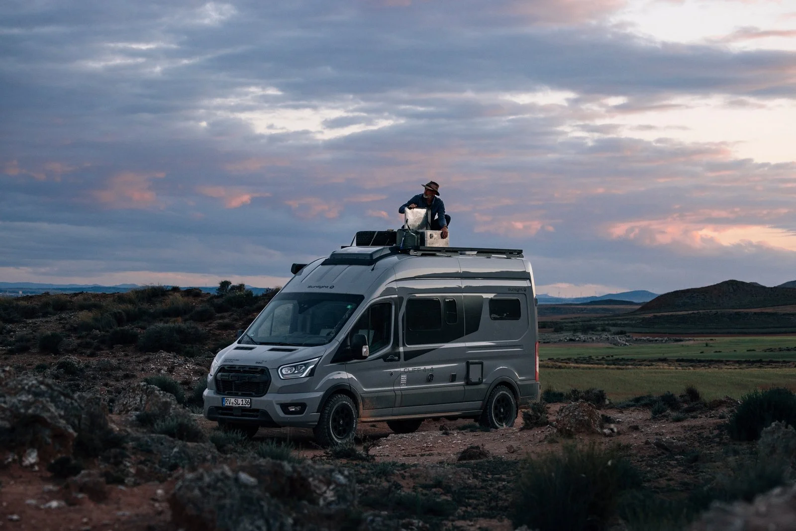Ein Wohnmobil ist in einer ländlichen Landschaft geparkt. Eine Person steht auf dem Dach des Fahrzeugs und arbeitet mit einer Ausrüstung. Der Himmel ist bewölkt und die Szenerie wirkt idyllisch und ruhig.