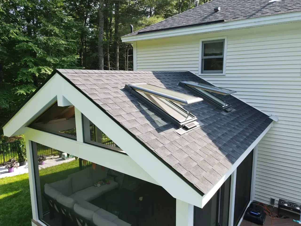 A house with a gabled roof featuring two skylights, and a screened porch with a white patio sofa inside, surrounded by trees and a grassy yard.