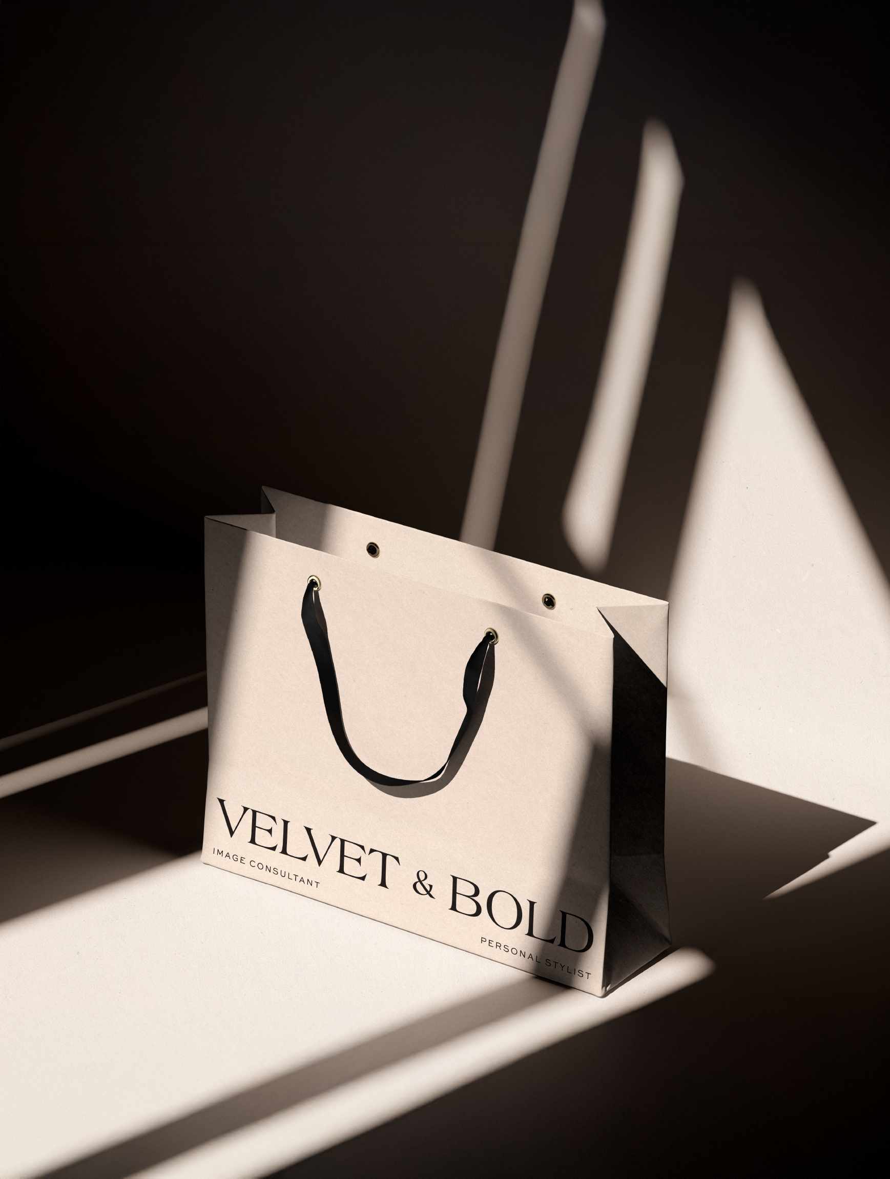 Sacola de papel bege com alças pretas, sob luz e sombras. Inscrição "Velvet & Bold" visível na sacola.