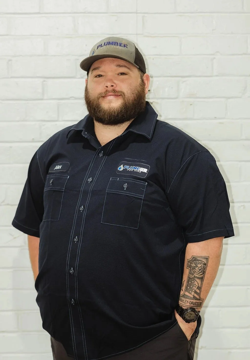 A man wearing a black shirt and cap with "Plumber" logo stands in front of a brick wall. He has a beard, visible tattoo on his forearm, and his hands in his pockets.