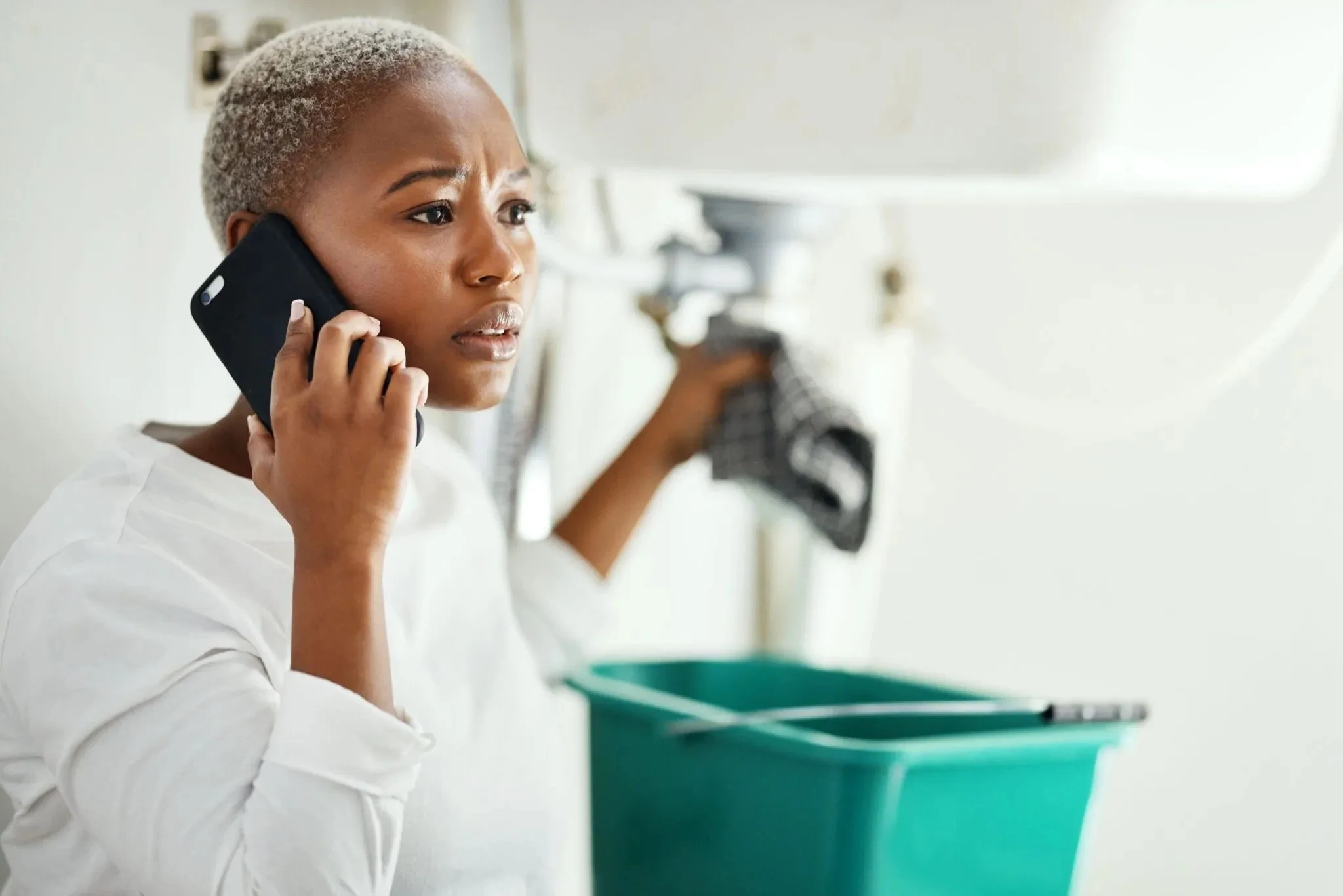 Woman on phone looking concerned, holding a dish towel, green bucket nearby.