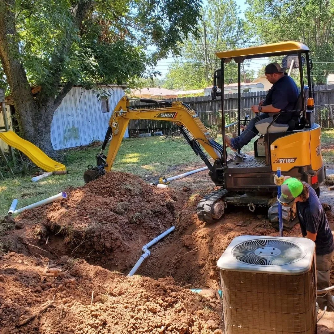 Construction site with a yellow mini excavator digging near PVC pipes, an outdoor air conditioning unit, and a person wearing a cap, in a backyard with trees and a yellow slide.