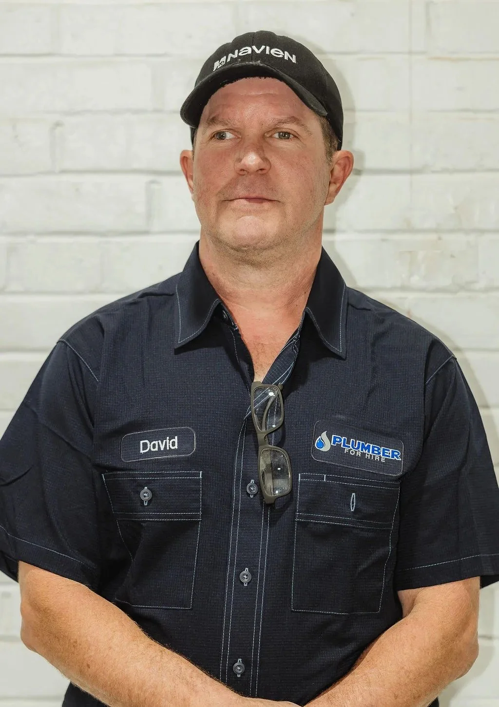 Man in a black cap and uniform shirt with name tag "David," standing against a white brick wall.