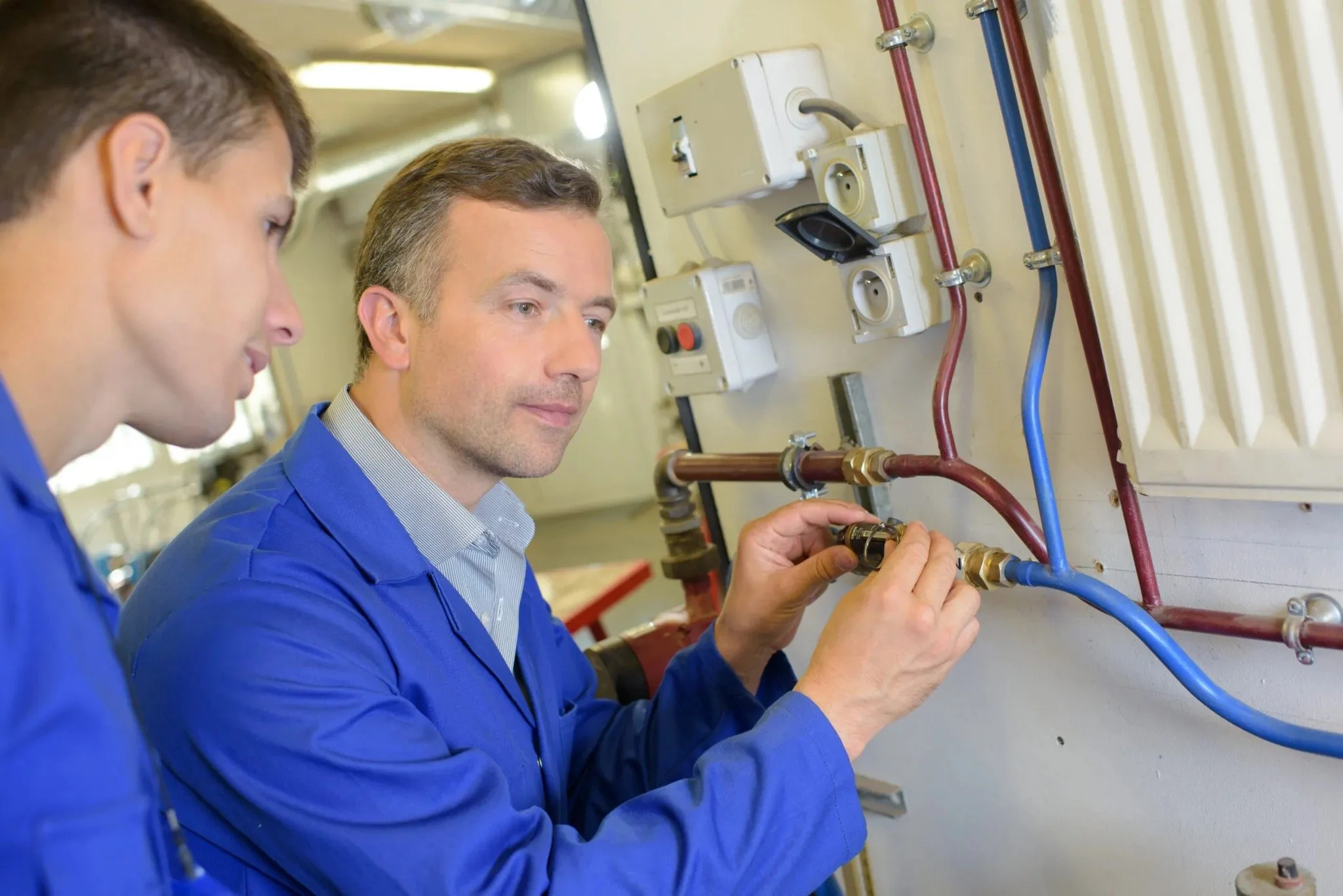 Two men in blue work uniforms examining plumbing pipes and fixtures on a wall.