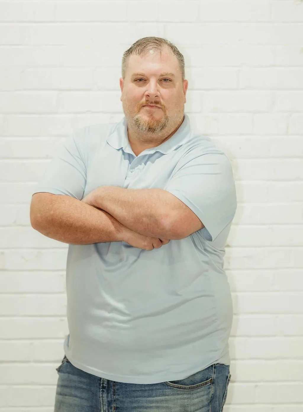 Man in light blue shirt and jeans standing with arms crossed against a white brick wall.