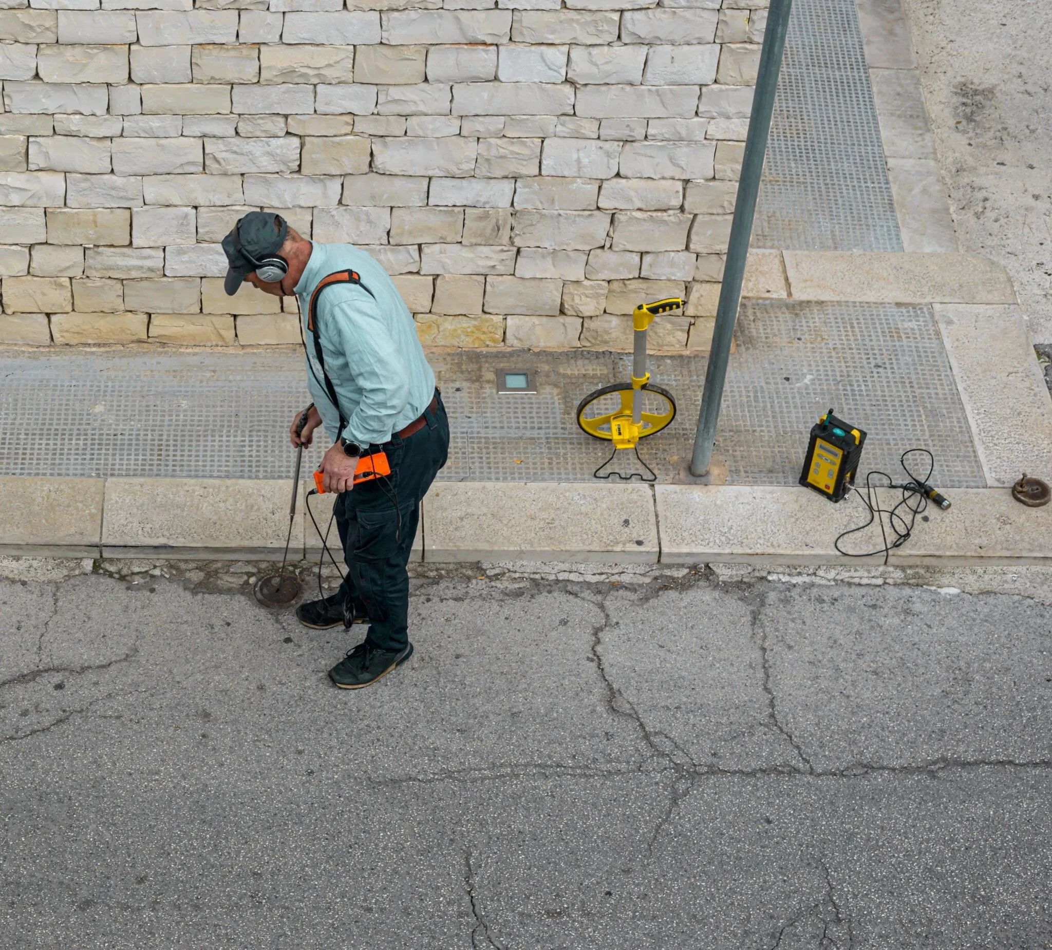 Person using leak detection equipment on a street next to a stone wall, wearing a cap and headphones, with tools and devices on the sidewalk.