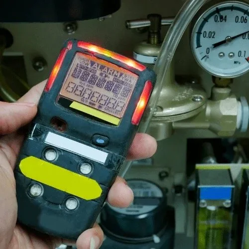 Person holding a handheld gas detector with a warning displayed on the screen, in front of industrial equipment with pressure gauges and valves.