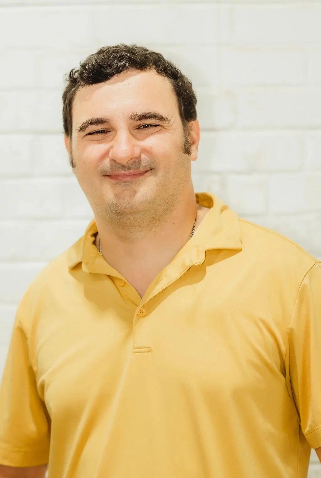 Smiling person wearing a yellow shirt in front of a white brick wall.