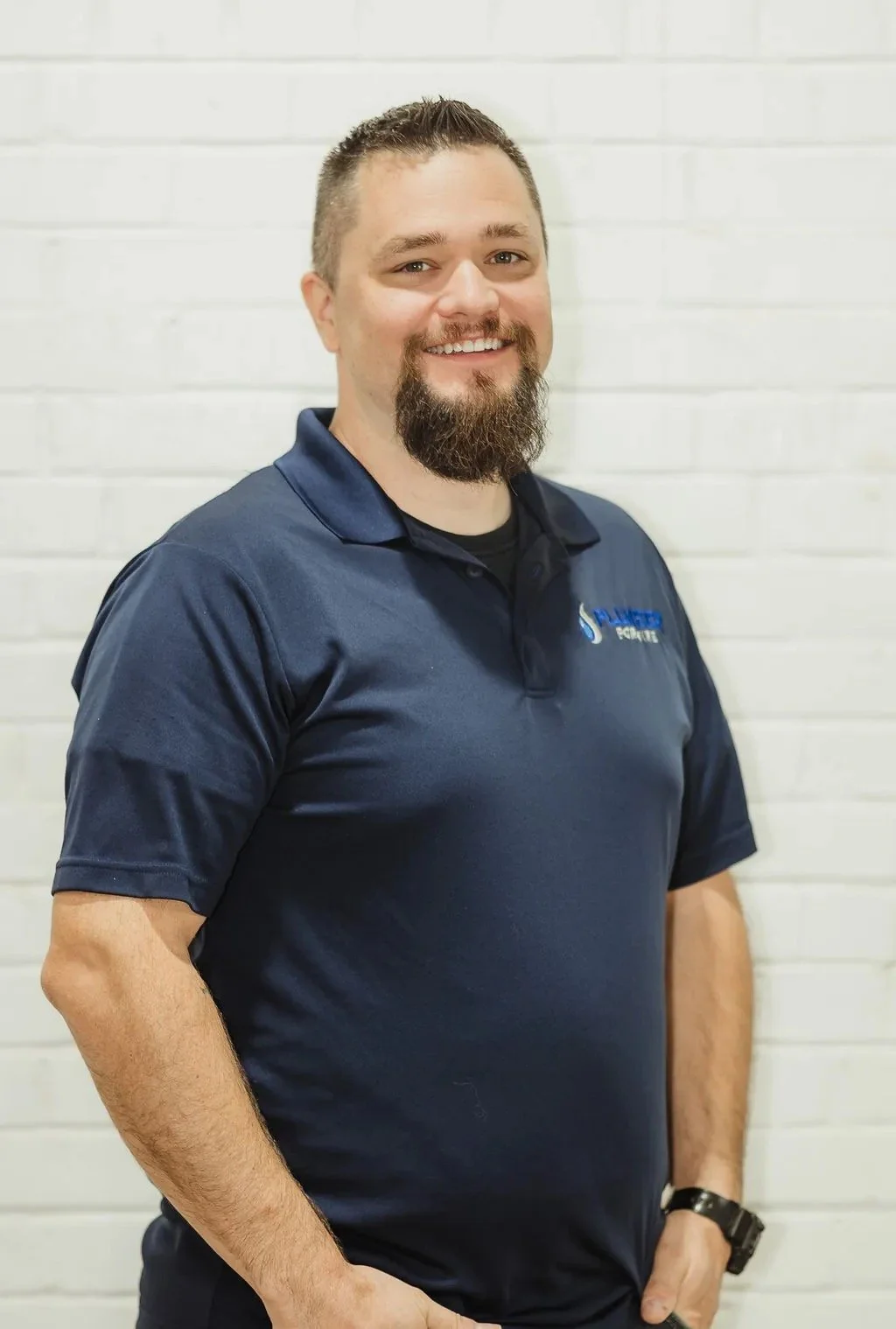 Smiling man with a beard wearing a navy blue polo shirt standing against a white brick wall.