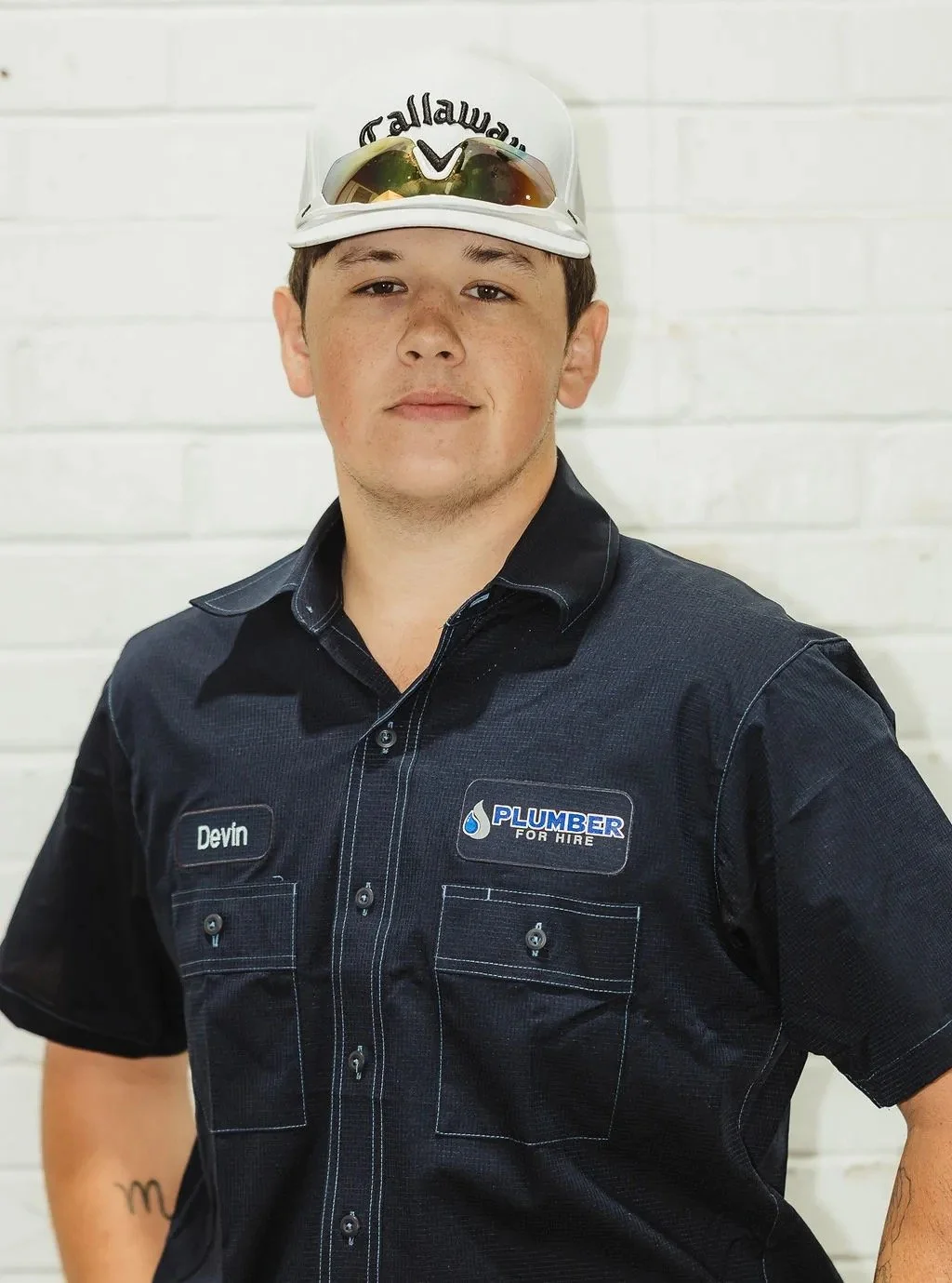 Young person wearing a Callaway hat and a dark uniform shirt with a "Plumber For Hire" logo, standing against a white brick wall.