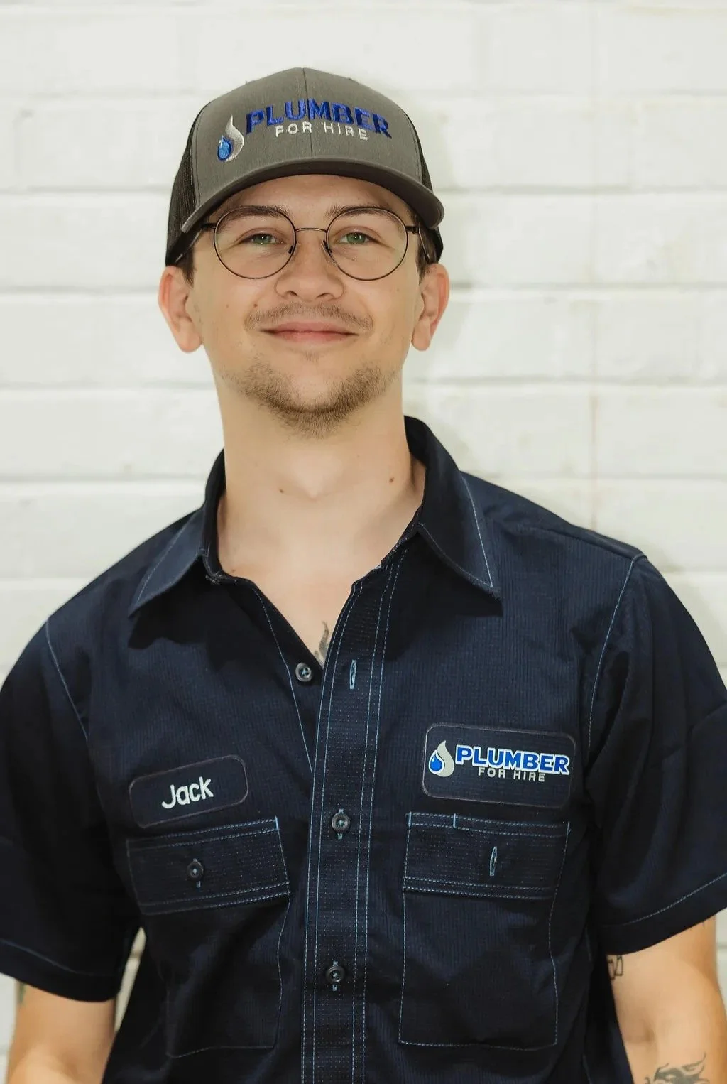 Person wearing a gray cap and blue uniform with "Plumber for Hire" logo, standing against a white brick wall.