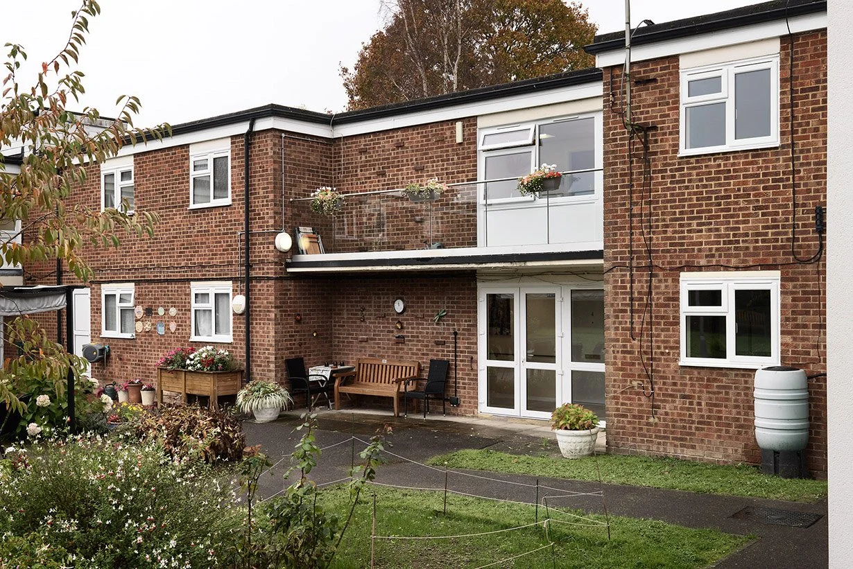 A two-story brick apartment building with a patio area featuring outdoor furniture, potted plants, and flower boxes on the balcony, surrounded by greenery and a garden.