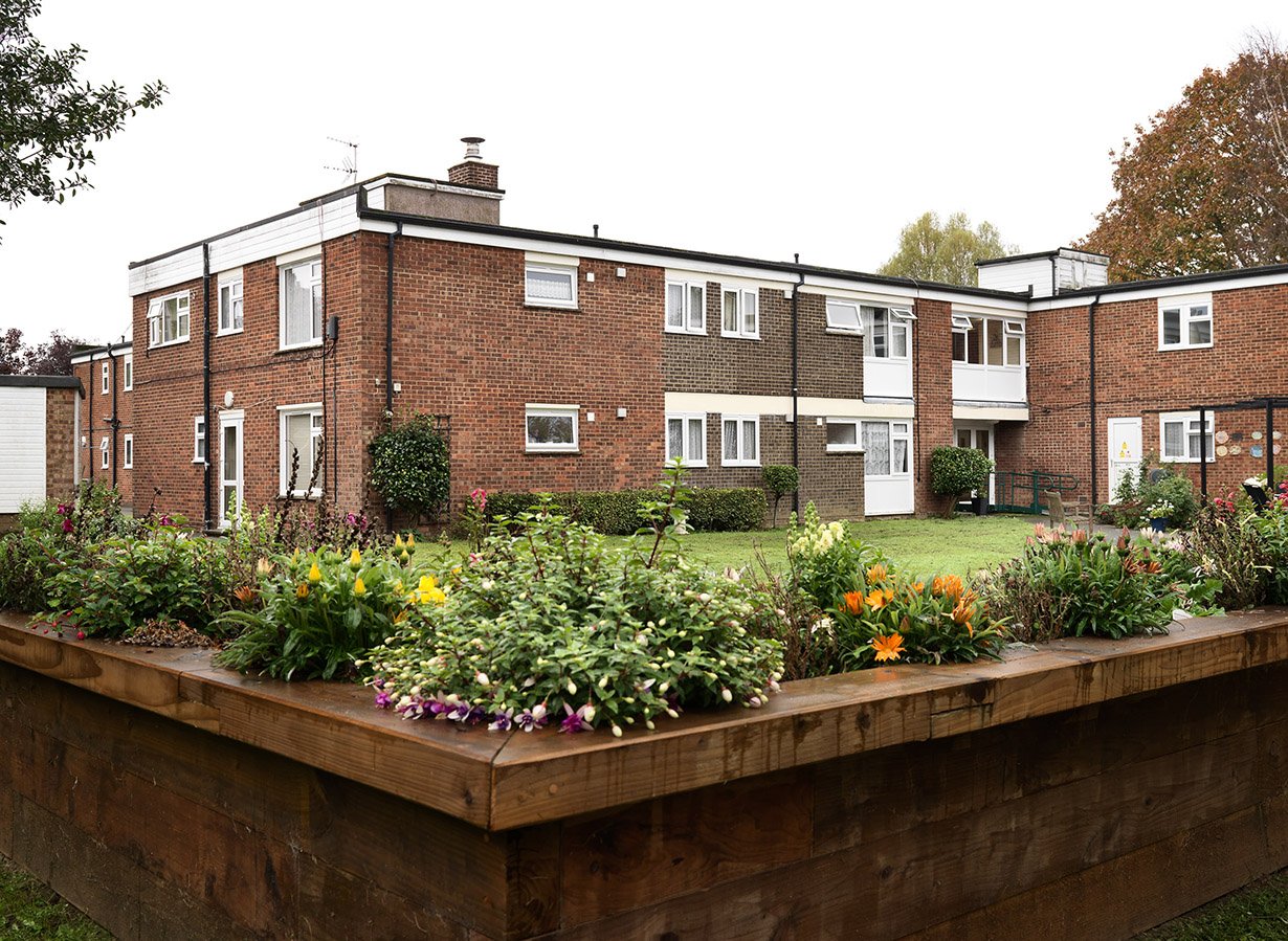 View of a multi-unit brick apartment complex with balconies and windows, surrounded by a green lawn and a flower garden with colorful flowers in the foreground.