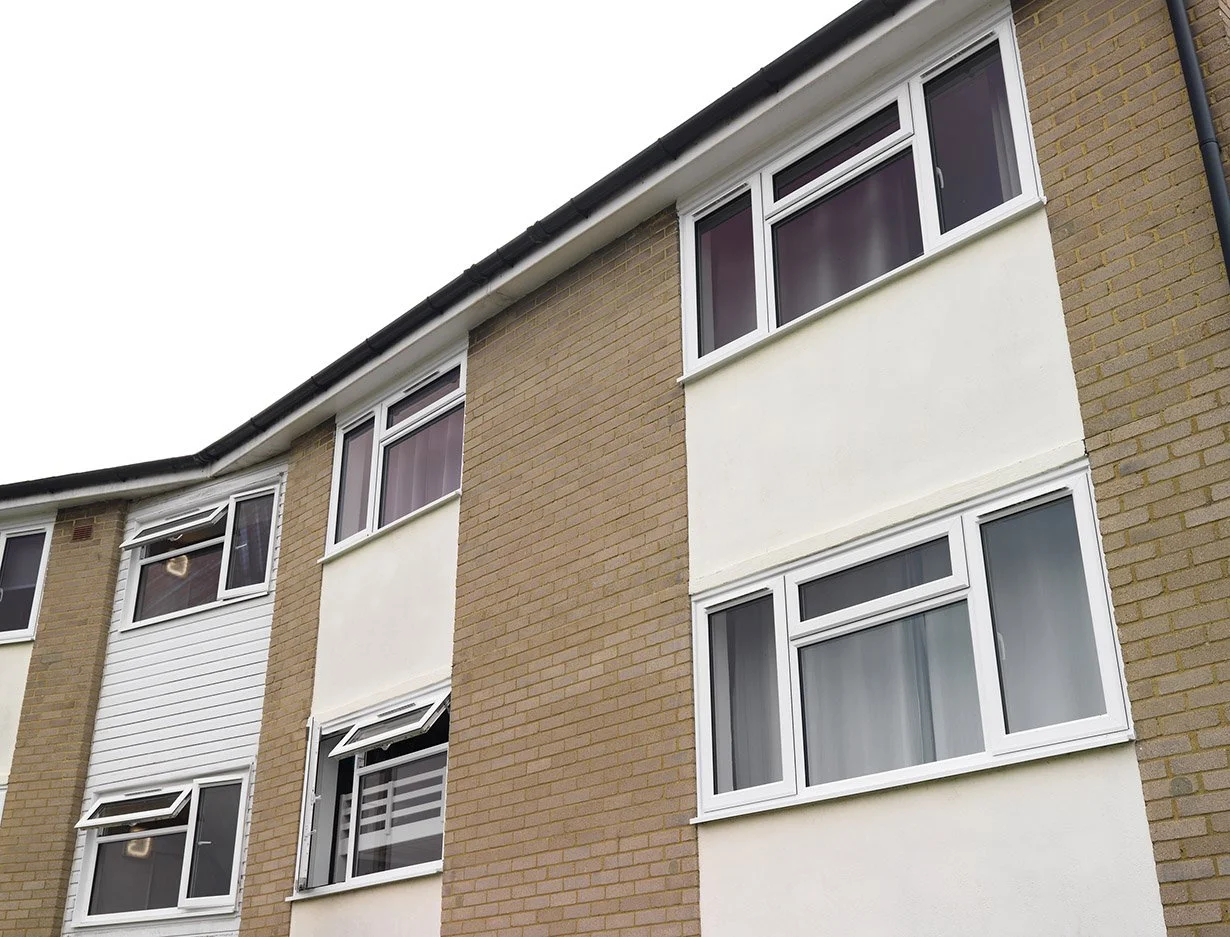 Close-up of a multi-story residential building with beige brick and white wall sections, multiple white-framed windows, some open, and a cloudy sky background.