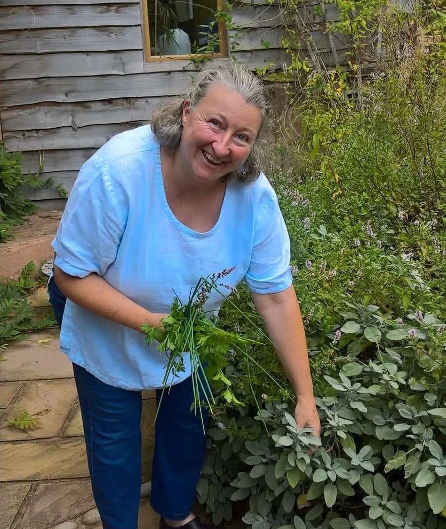 A smiling woman with gray hair wearing a light blue shirt and dark pants, picking herbs or plants in a garden next to a wooden house with a small window, surrounded by green foliage.
