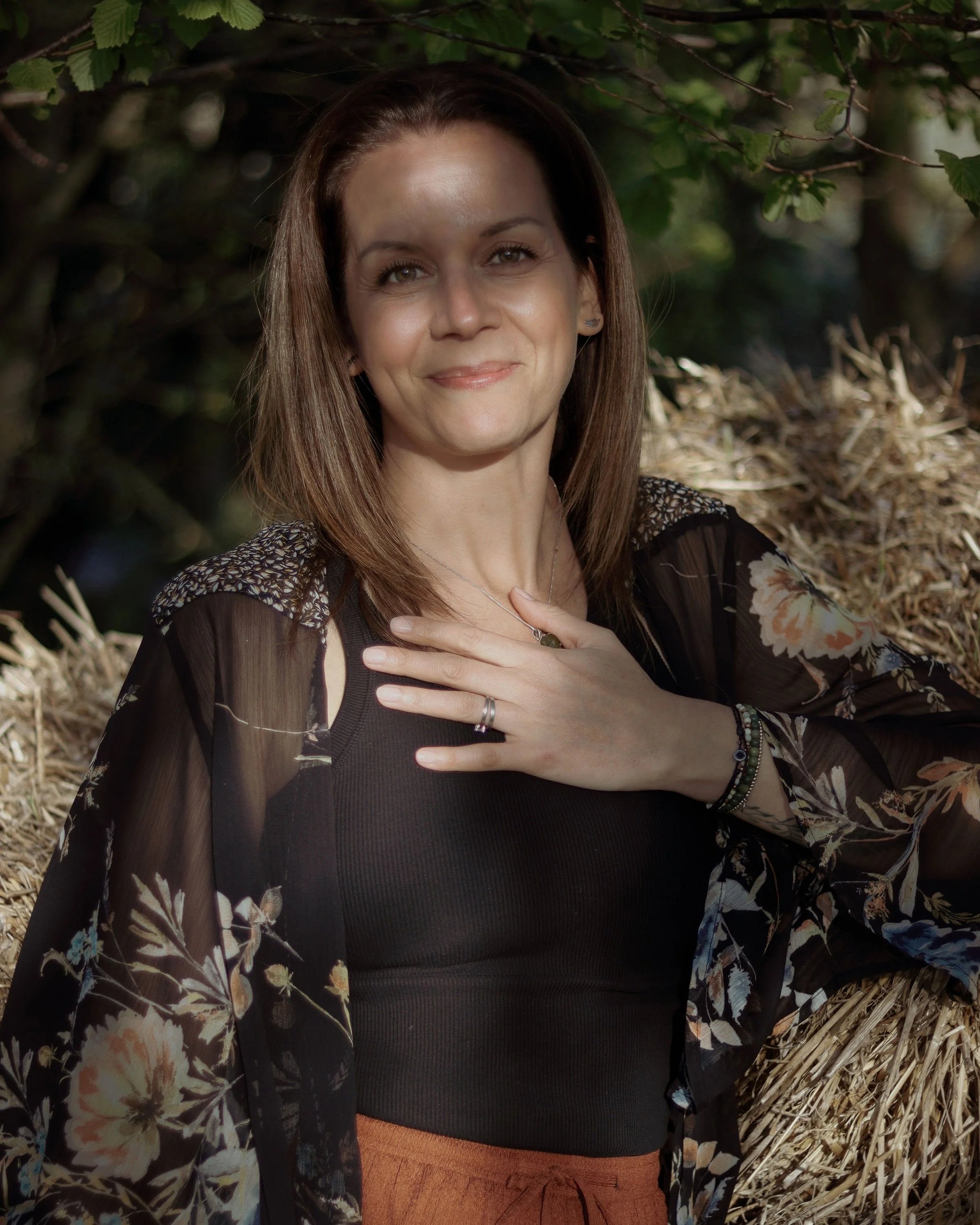 A woman with brown hair sitting outdoors in front of hay bales surrounded by green foliage, smiling with one hand on her chest.